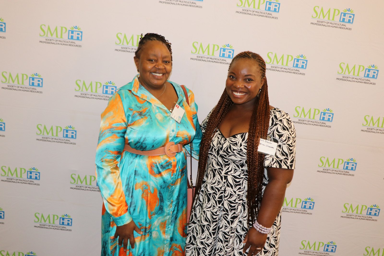 Two women in professional attire smiling at camera at SMPHR-branded photo backdrop