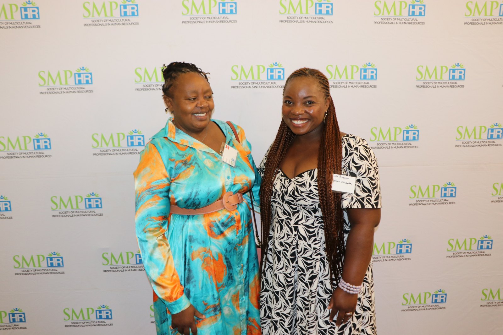 Two women in professional attire smiling at camera at SMPHR-branded photo backdrop