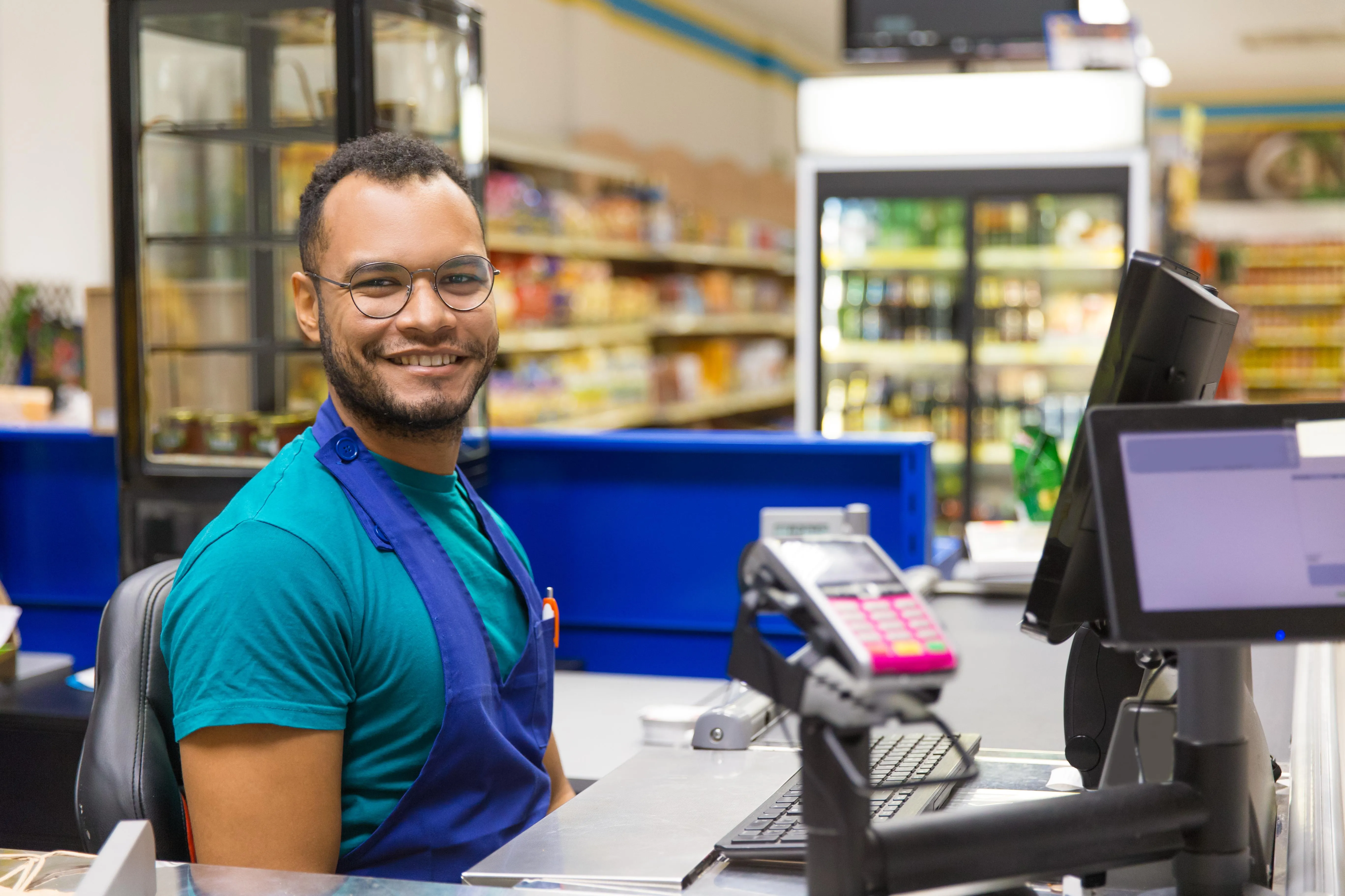 Caissier souriant avec lunettes et tablier bleu, assis à une caisse de supermarché.