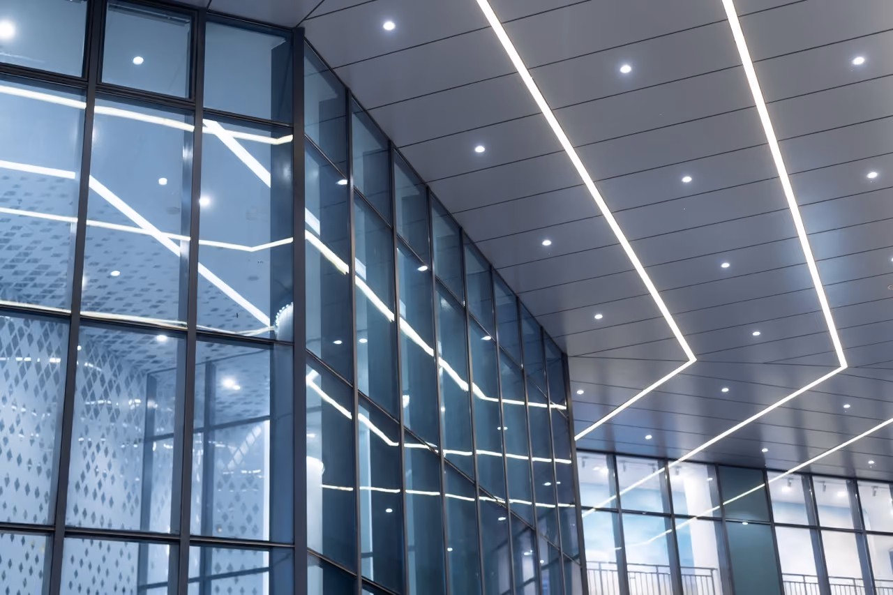 Moderne glazen gevel van een gebouw met reflecties van witte lijnverlichting aan het plafond binnenin.