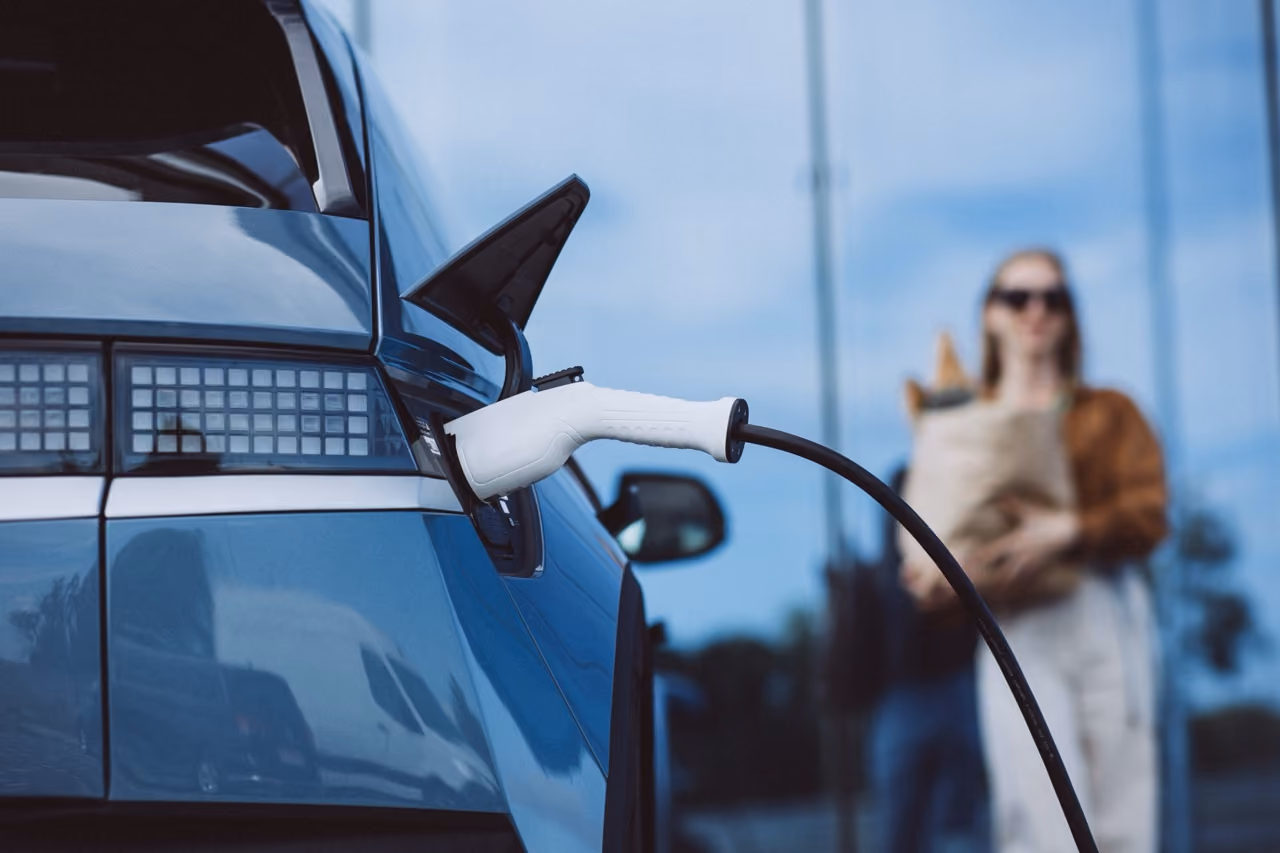Een blauw elektrisch voertuig wordt opgeladen met een wit oplaadpunt, op de achtergrond staat een vrouw met een boodschappentas.