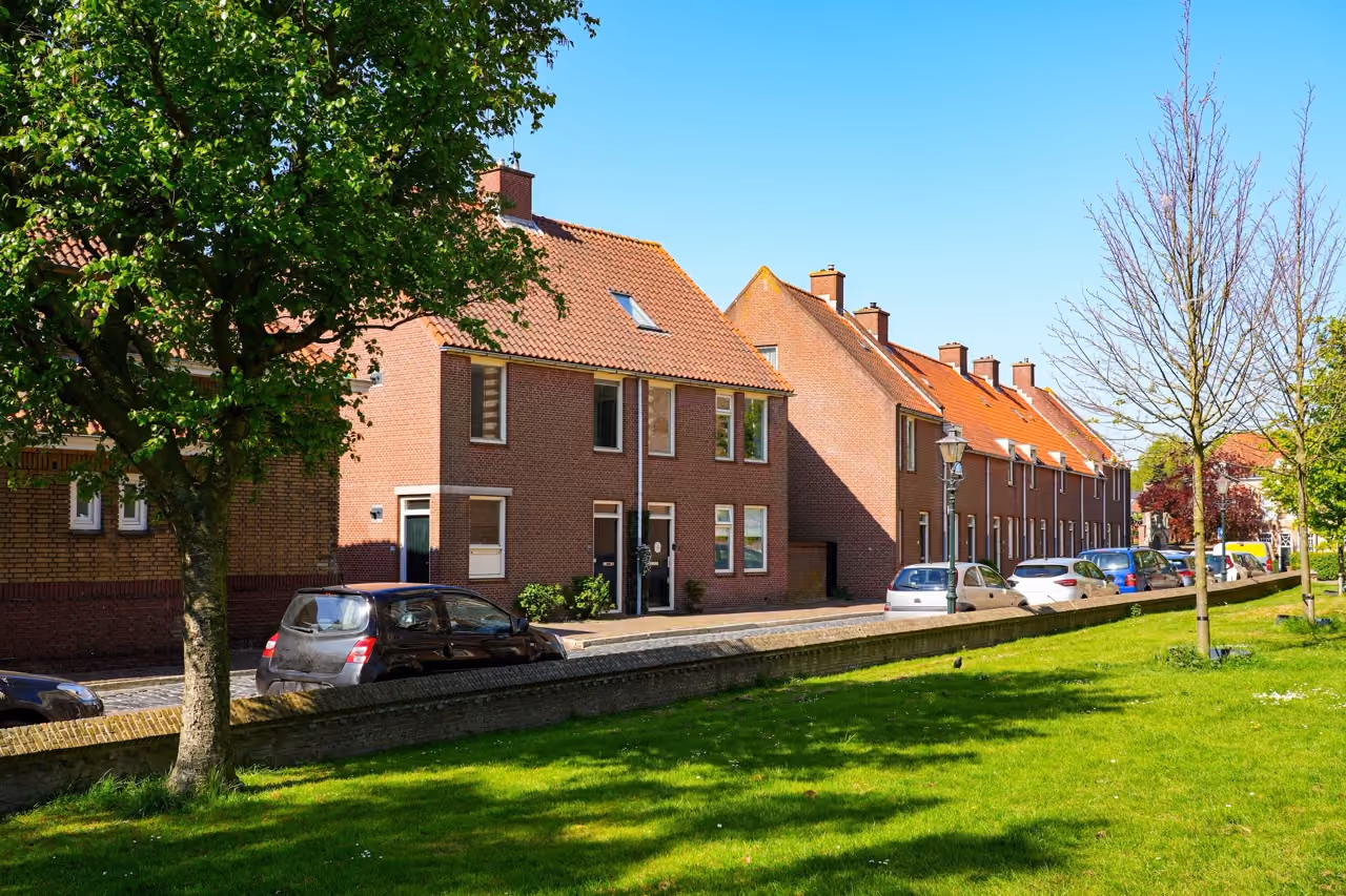 Rij van traditionele Nederlandse bakstenen huizen met rode daken langs een straat met geparkeerde auto's, groene grasvelden en bomen onder een heldere blauwe lucht.
