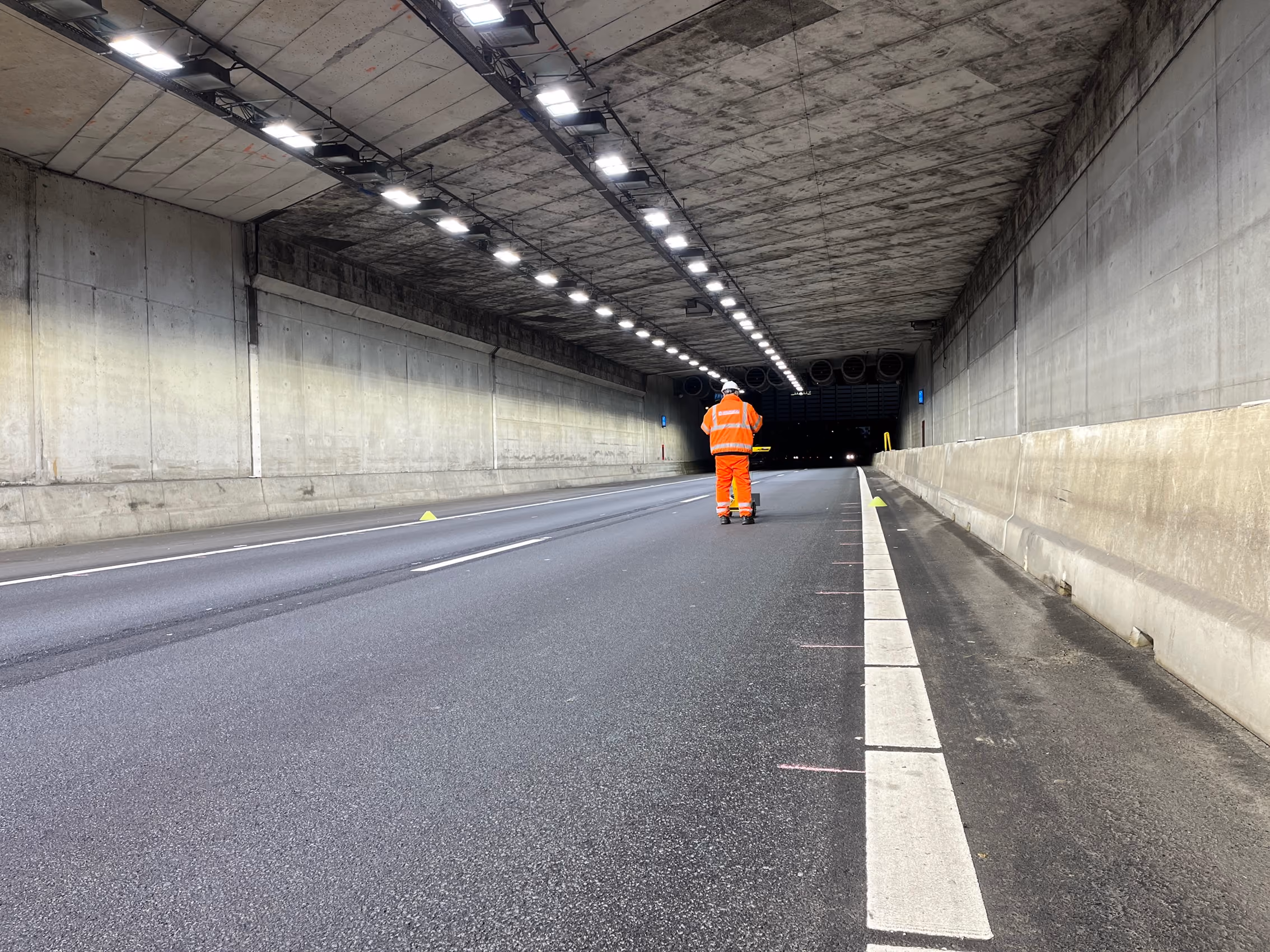 Een persoon in een oranje veiligheidsoutfit en helm loopt in een brede betonnen tunnel met verlichtingsarmaturen aan het plafond en een glad wegdek.