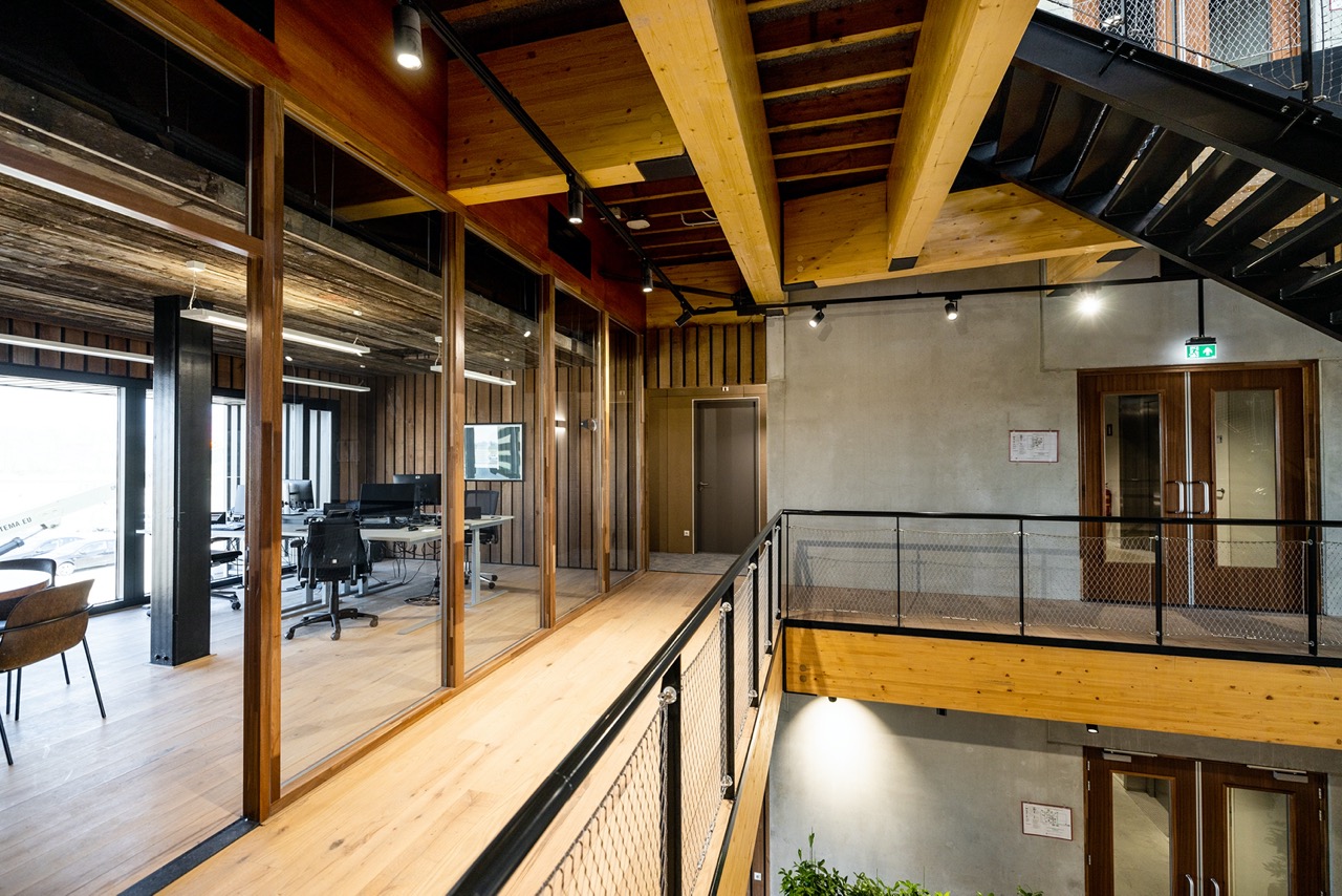 Moderne kantoorruimte met glazen wanden en houten vloer, zicht op bureaus met computers en stoelen, houten balken aan het plafond en metalen trap.