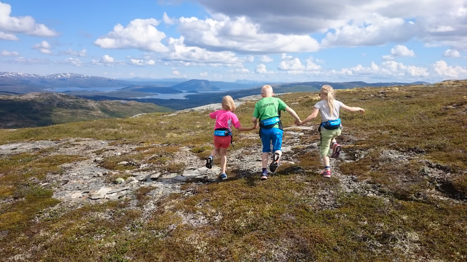 Tre barn løper hånd i hånd over steinete, mosegrodd terreng med fjell og innsjø i bakgrunnen.