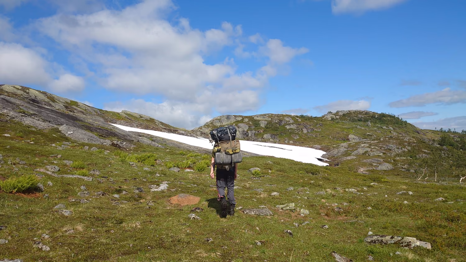 En person med stor ryggsekk går på steinete, grønt fjellterreng med snøflekk under blå himmel.
