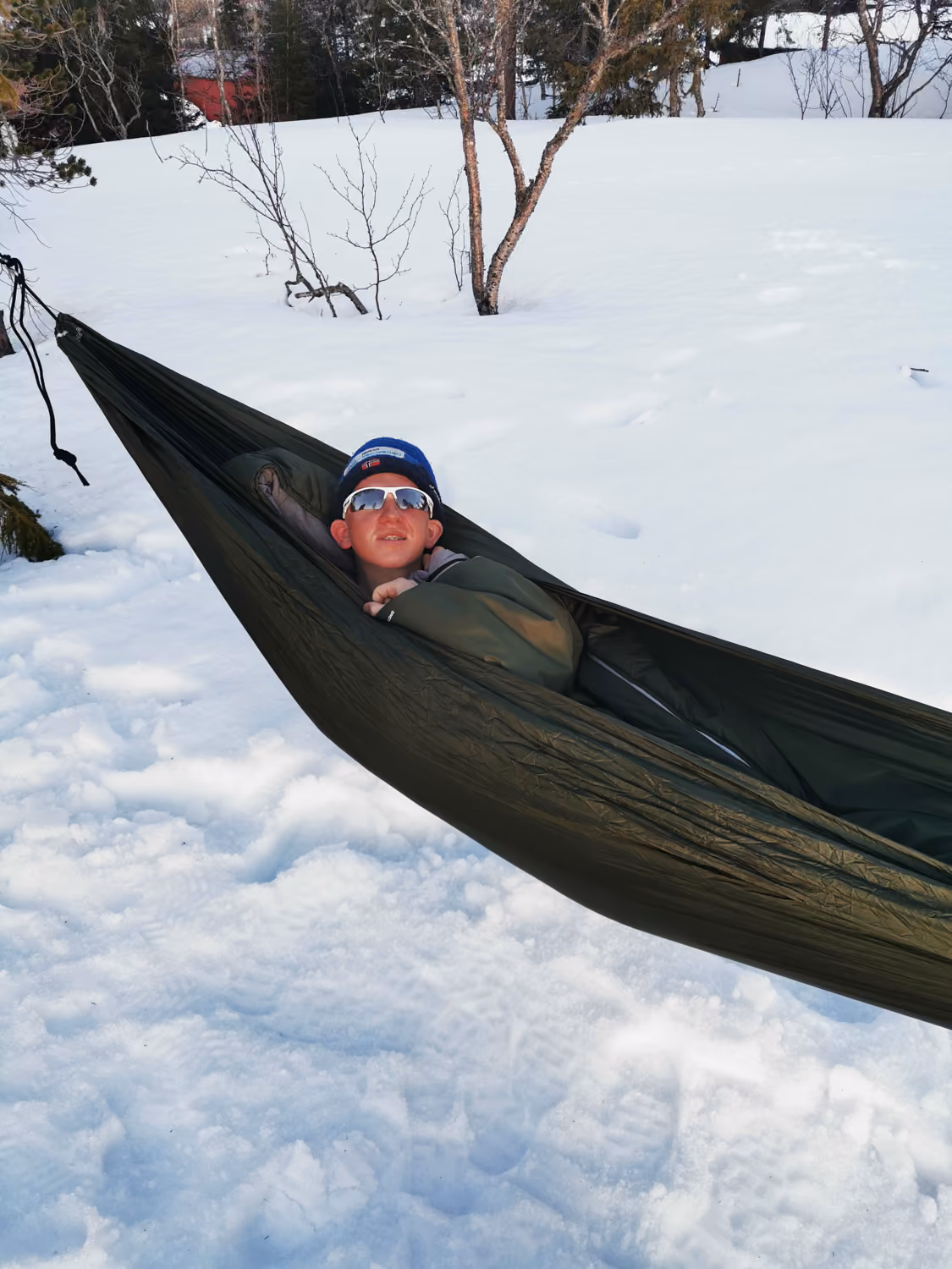 Person med blå lue og solbriller som ligger i hengekøye over snødekt mark.