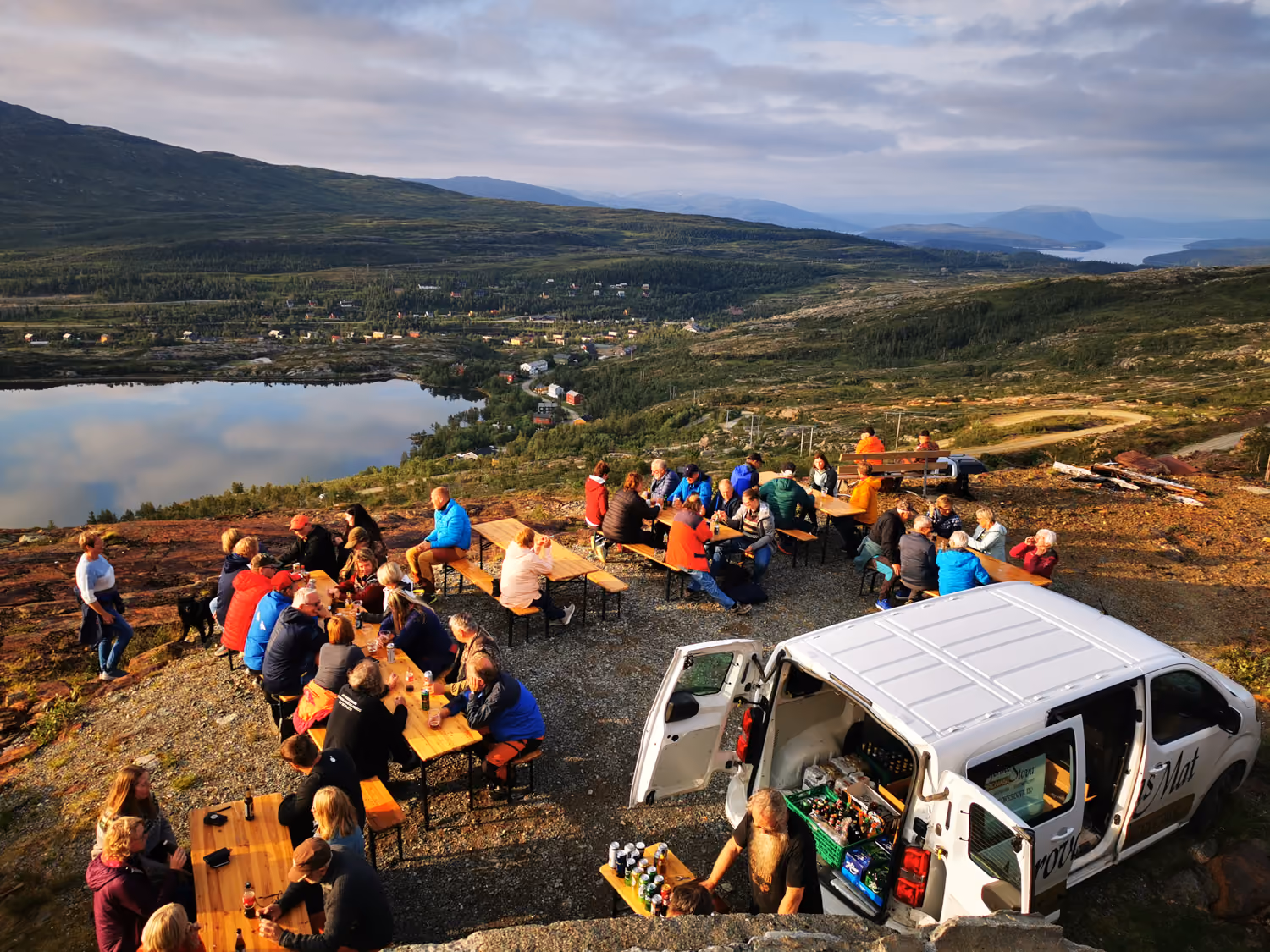 Folk samles utendørs rundt bord nær en hvit varebil med fjell, innsjø og landsby i bakgrunnen.