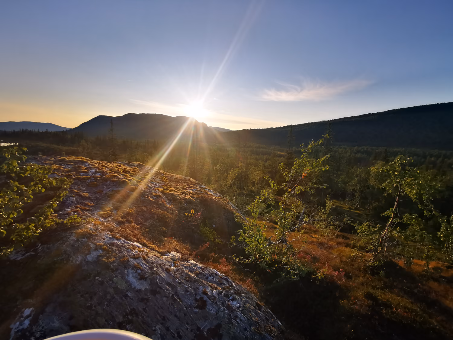 Solen går ned bak fjell med skog og steiner i forgrunnen under en klar himmel.