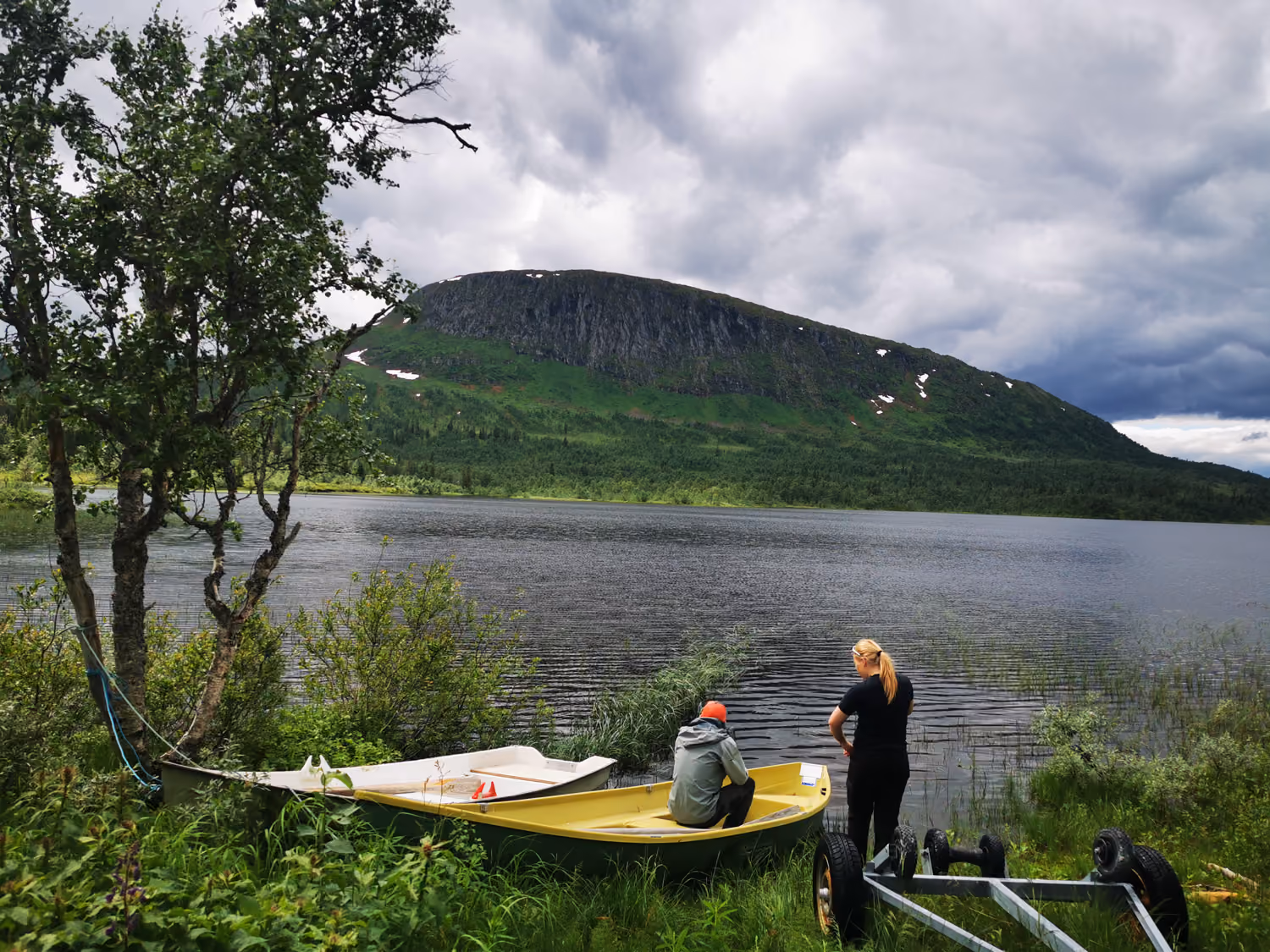 To personer ved en innsjø med to båter og et fjell i bakgrunnen under overskyet himmel.