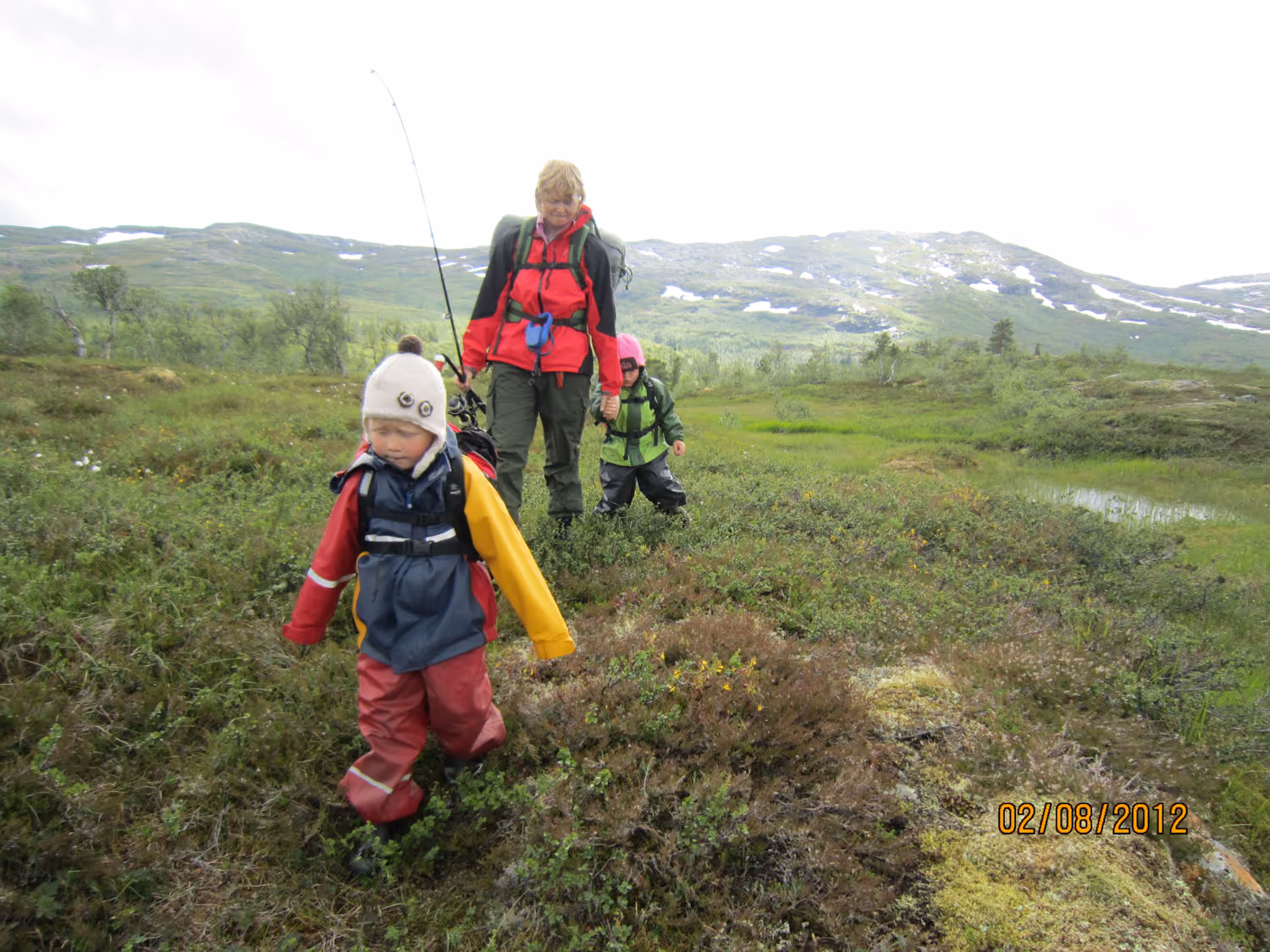 To barn og en voksen som går med fiskeutstyr i et grønt fjellandskap.