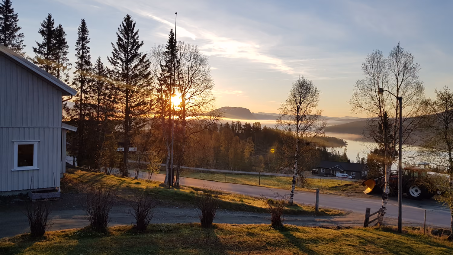 Solnedgang bak trær og hus med utsikt mot innsjø, fjell og traktor ved en landevei.