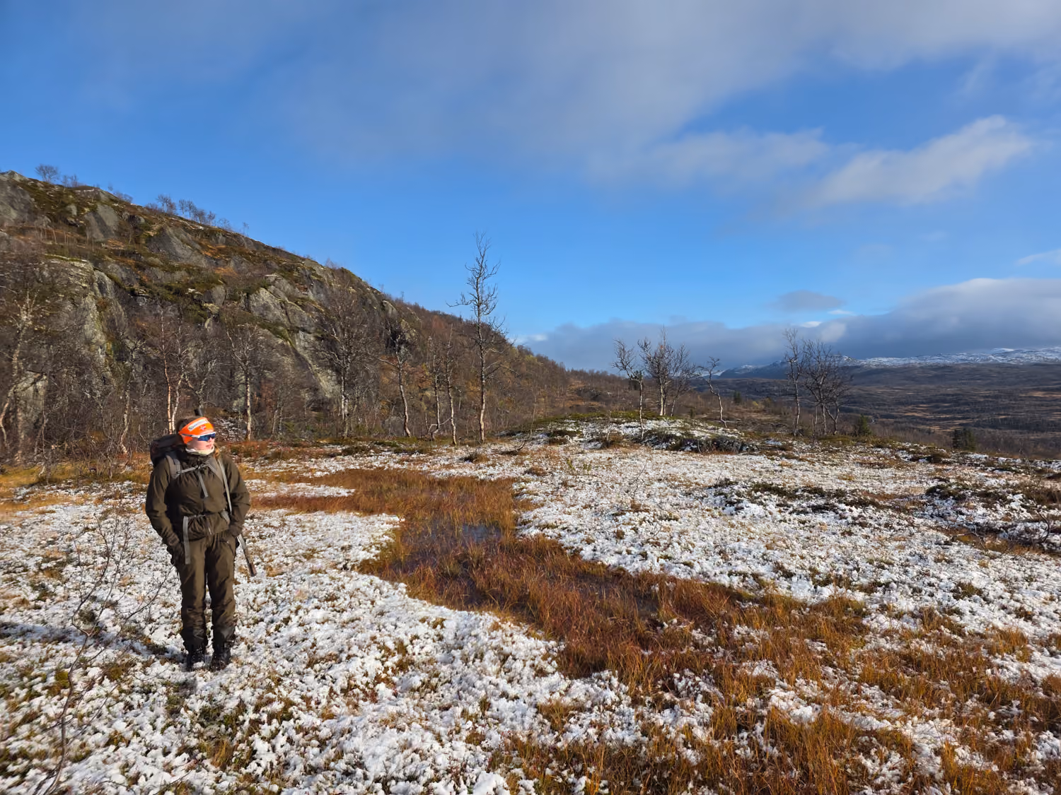 En person med oransje lue står i et snødekket, åpent terreng med fjell og trær i bakgrunnen.
