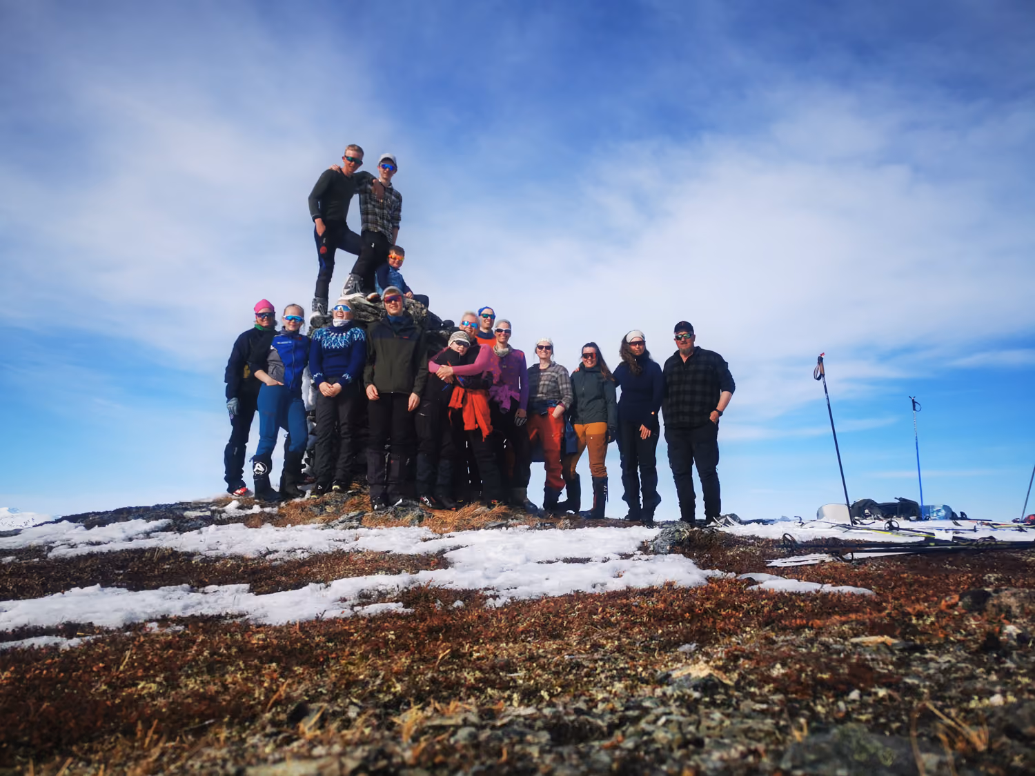 Gruppe på 15 mennesker på topp av fjell med snø og skistaver under blå himmel.