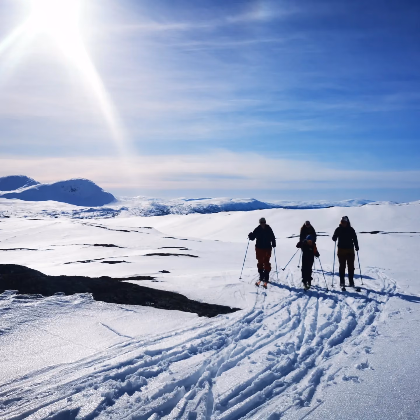 Tre personer på langrenn går gjennom snødekte fjell under en solrik, blå himmel.