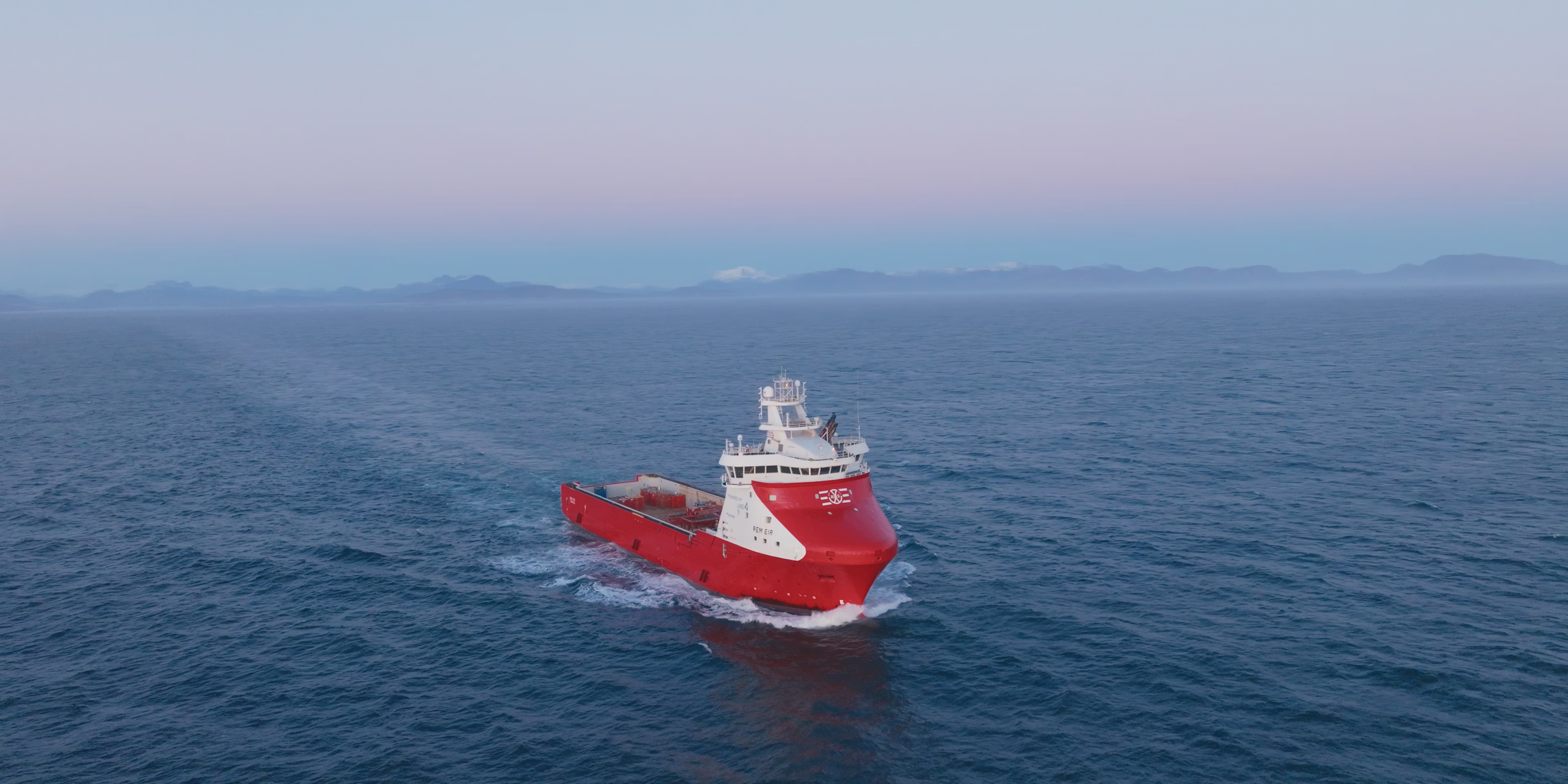 Red and white ship sailing on calm blue ocean with distant mountains under clear sky.