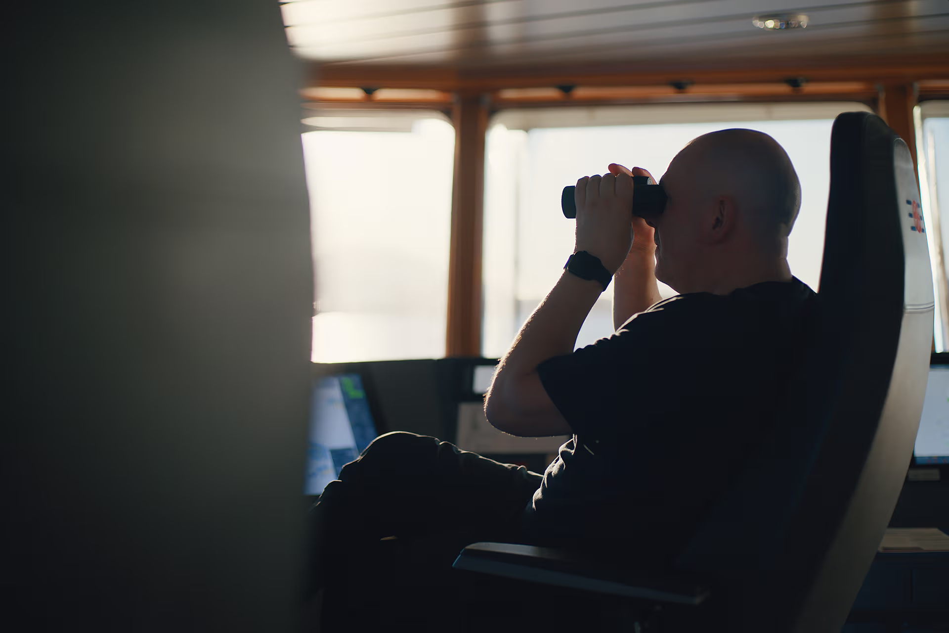 Man sitting on a chair inside a ship's cabin looking through binoculars toward the window.