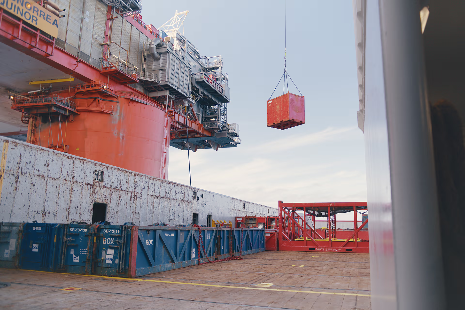 Red container being lifted by crane on industrial dock with stacked blue containers nearby.