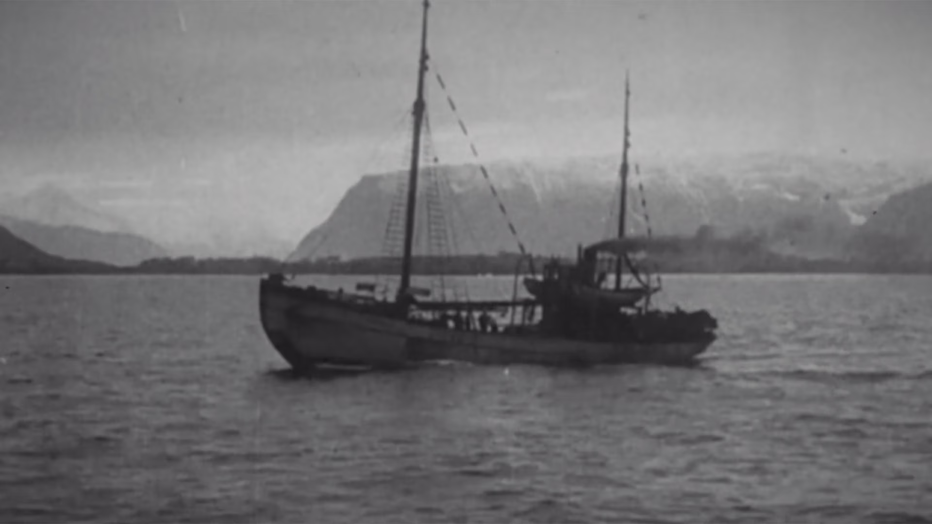 Black and white photo of a two-masted fishing boat on water with mountains in the background.