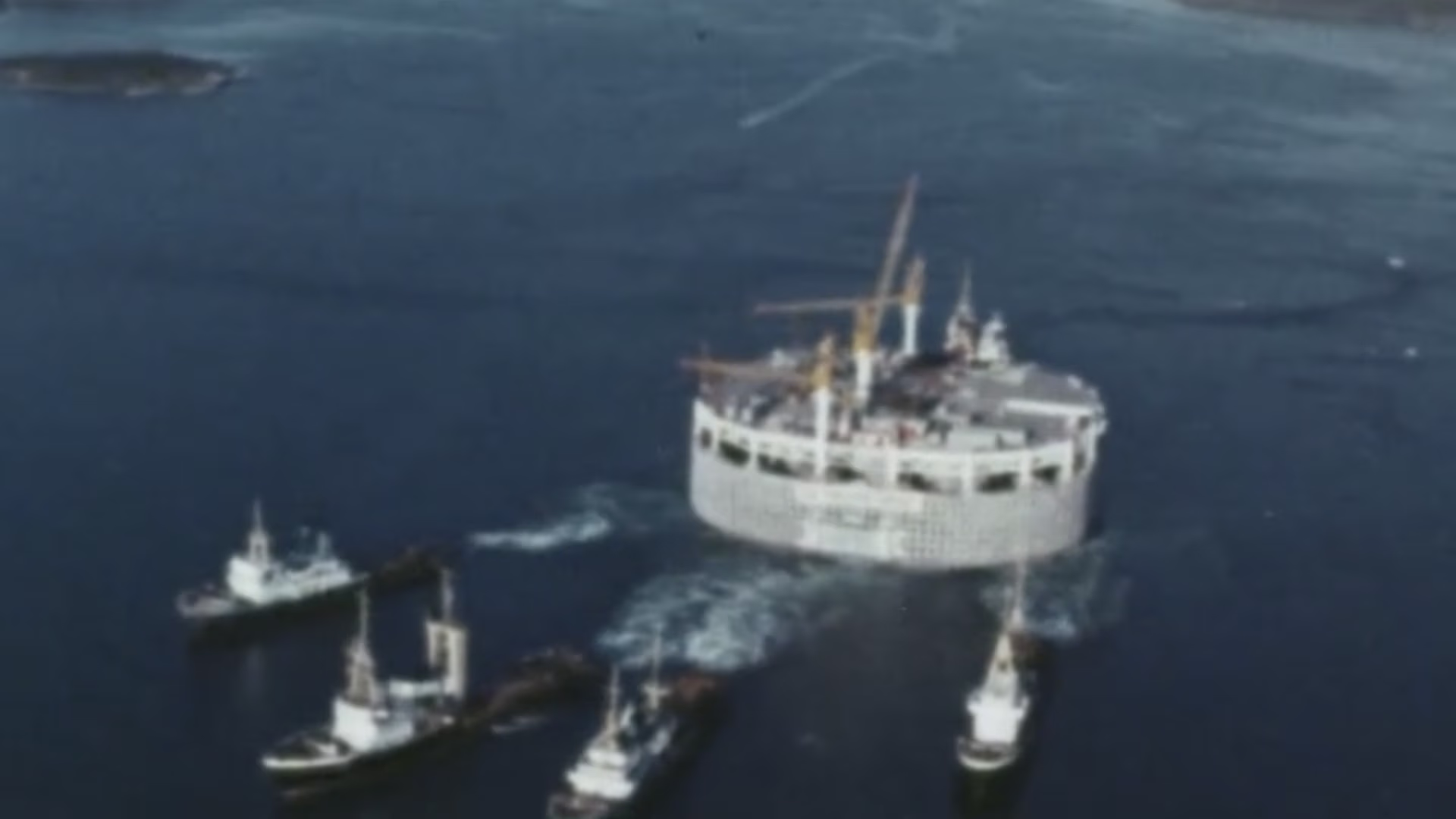 Large circular offshore platform being towed by several tugboats on the ocean.
