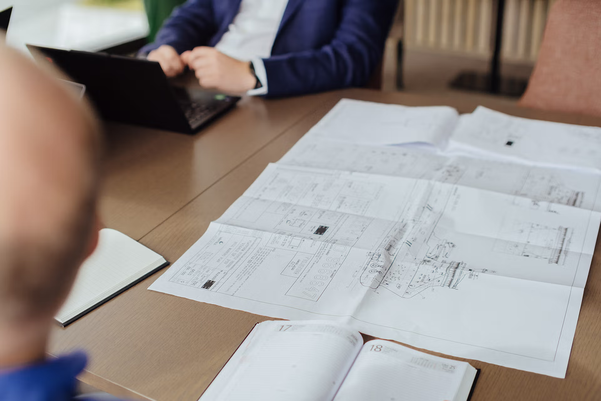 Table with blueprints, open notebook, and two people working, one on laptop.