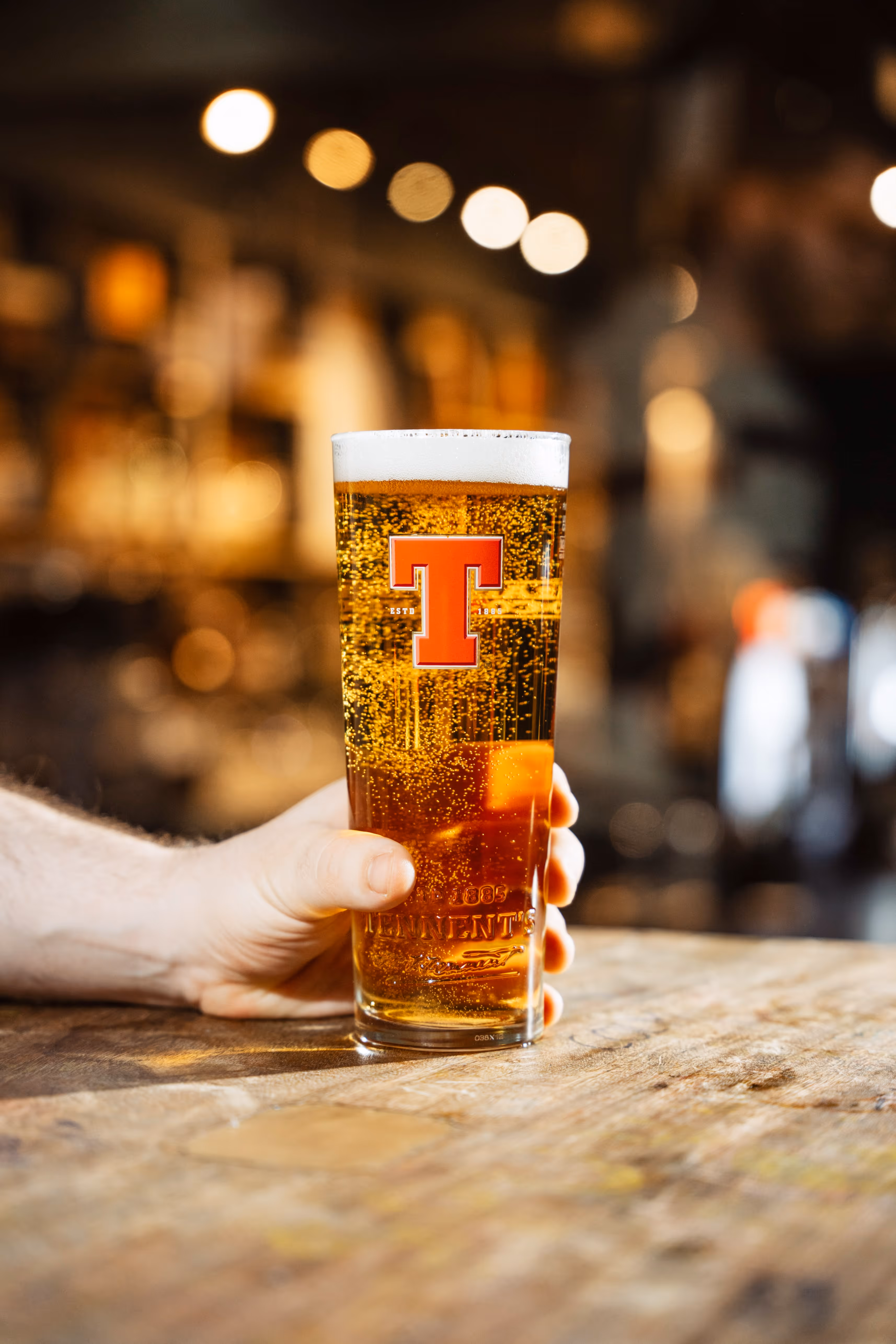 A hand holding a pint of Tennent's on a bar table