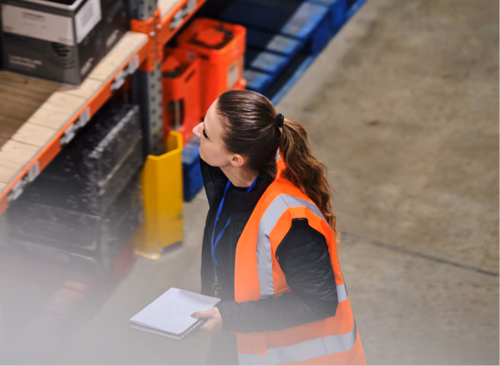 Woman in orange safety vest holding notebook, inspecting shelves in a warehouse.