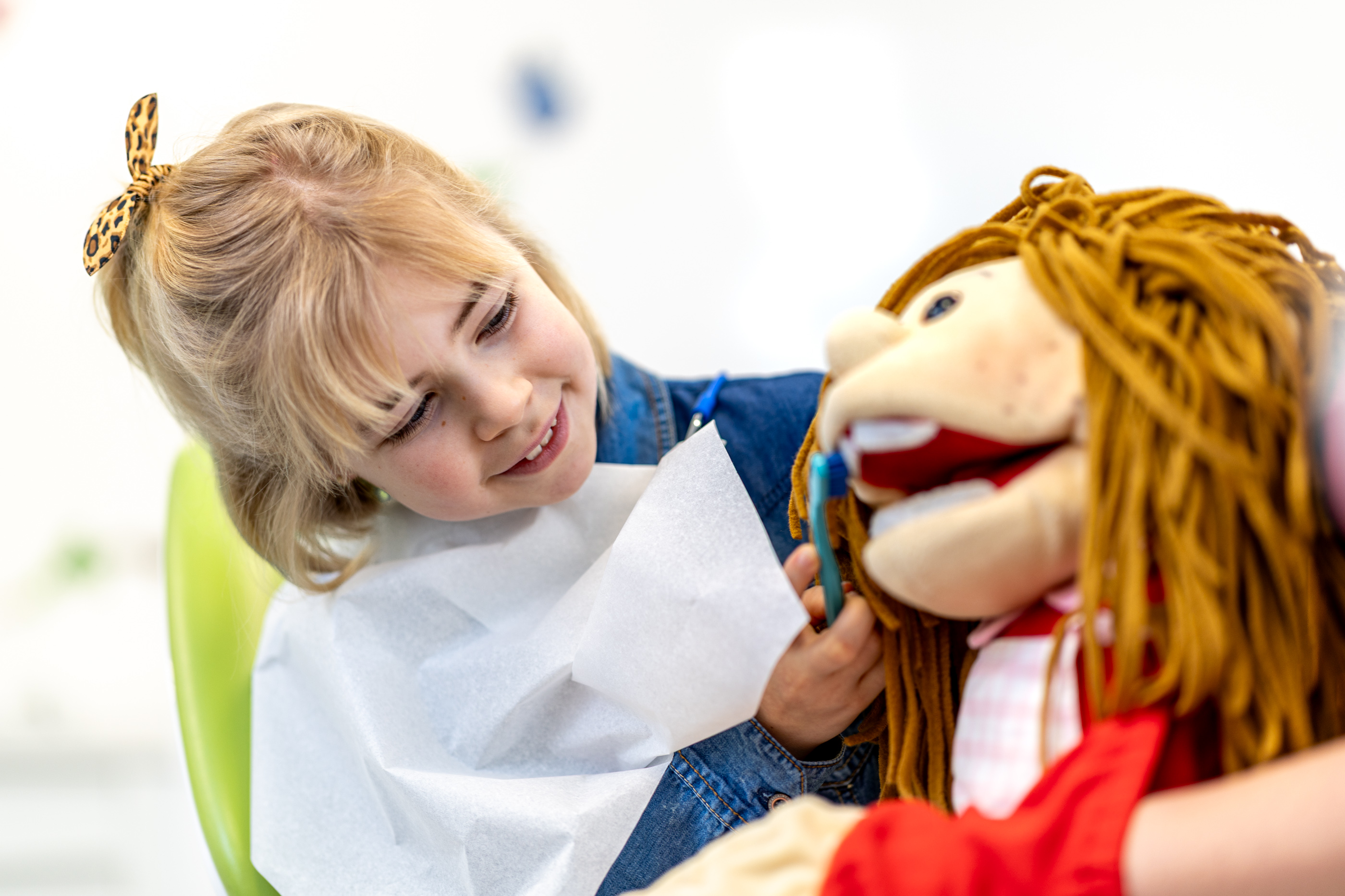 Ein kleines Mädchen mit einem Zahnputzlätzchen lächelt und putzt die Zähne einer großen Puppe mit braunen Wollhaaren.