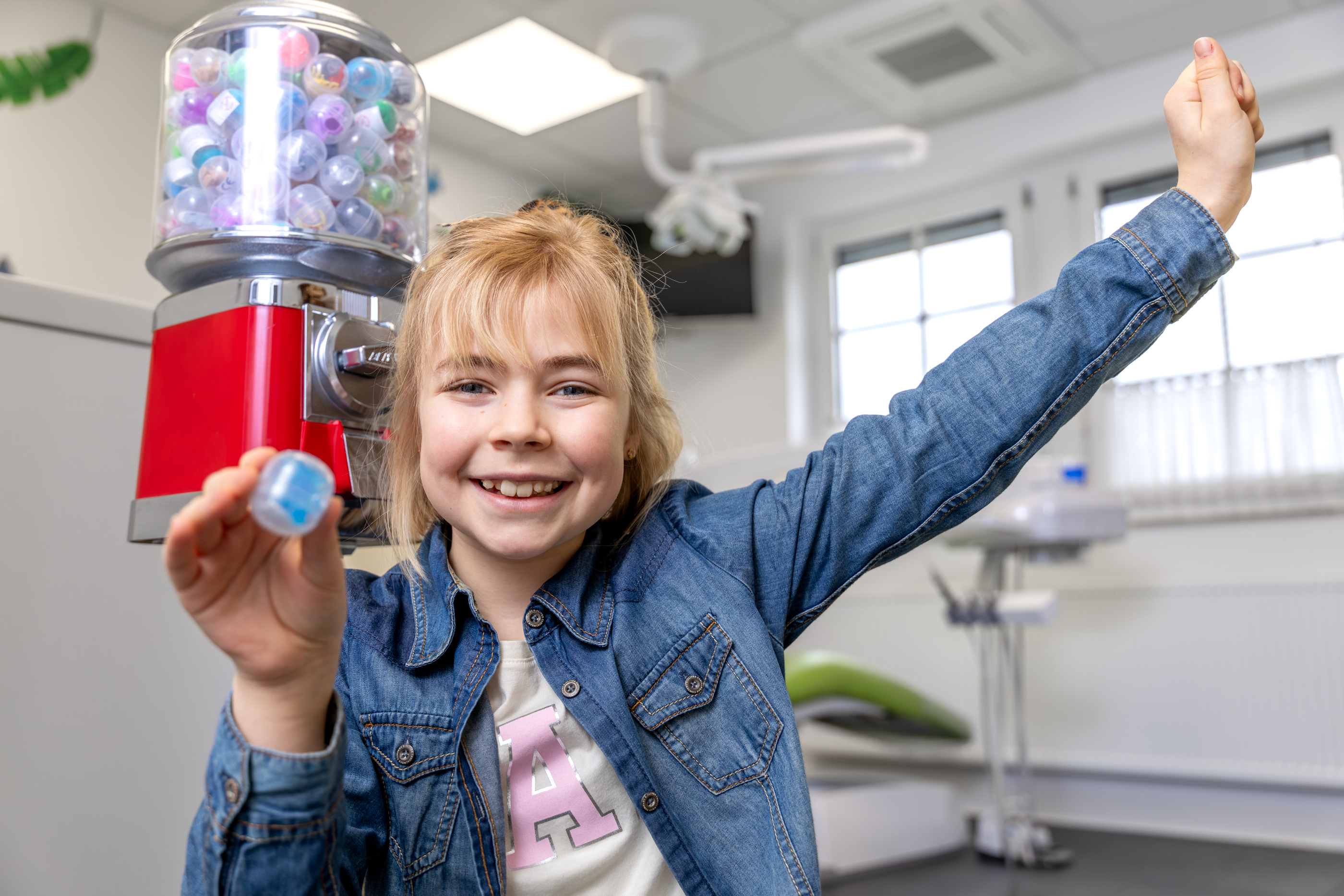 Lächelndes Mädchen in einer Zahnarztpraxis, das ein blaues Kapsel-Spielzeug in der Hand hält, im Hintergrund ein Spielzeugautomat.
