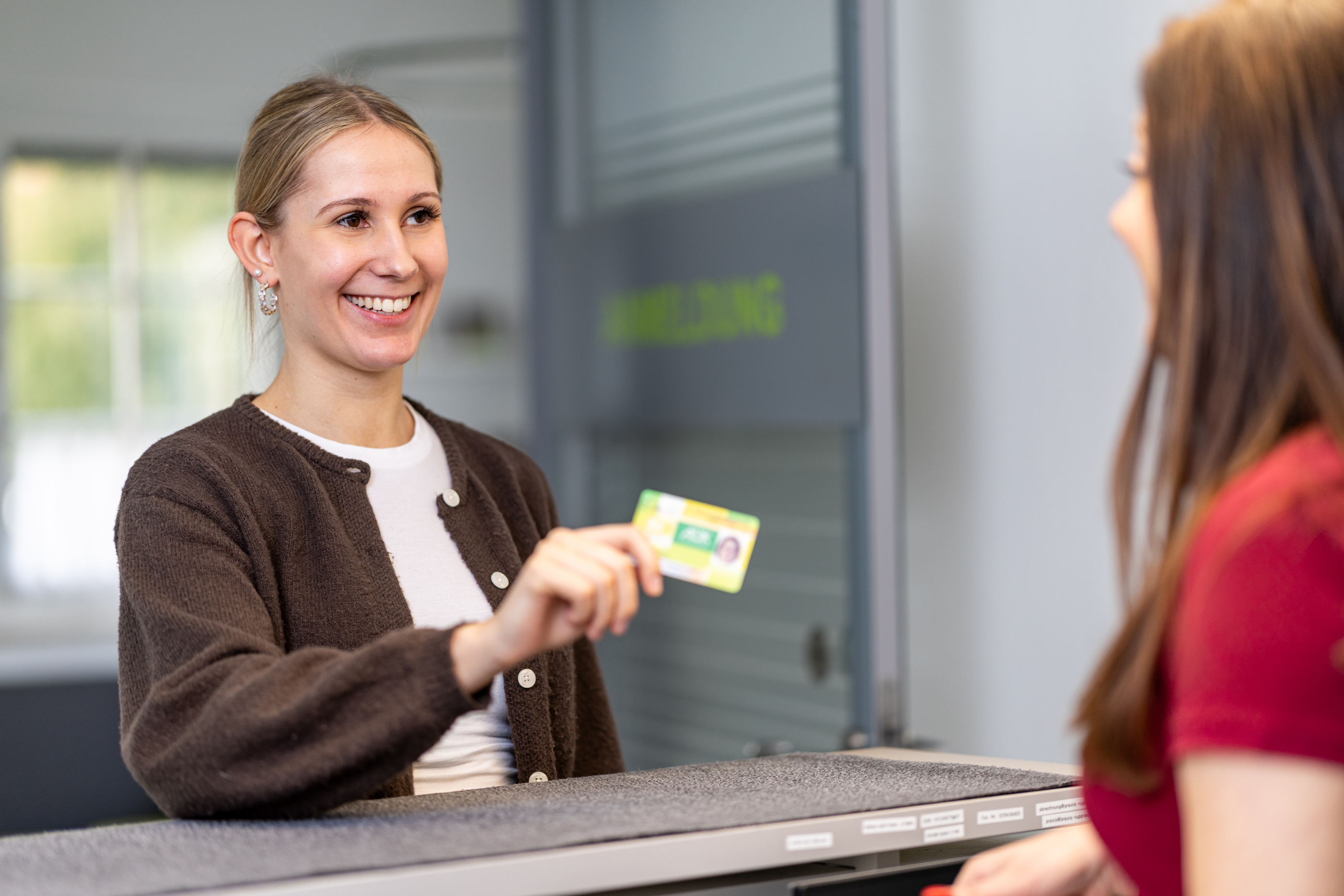Eine lächelnde Frau, die an einem Empfangstresen einen Personalausweis in der Hand hält und sich mit einer anderen Person unterhält.