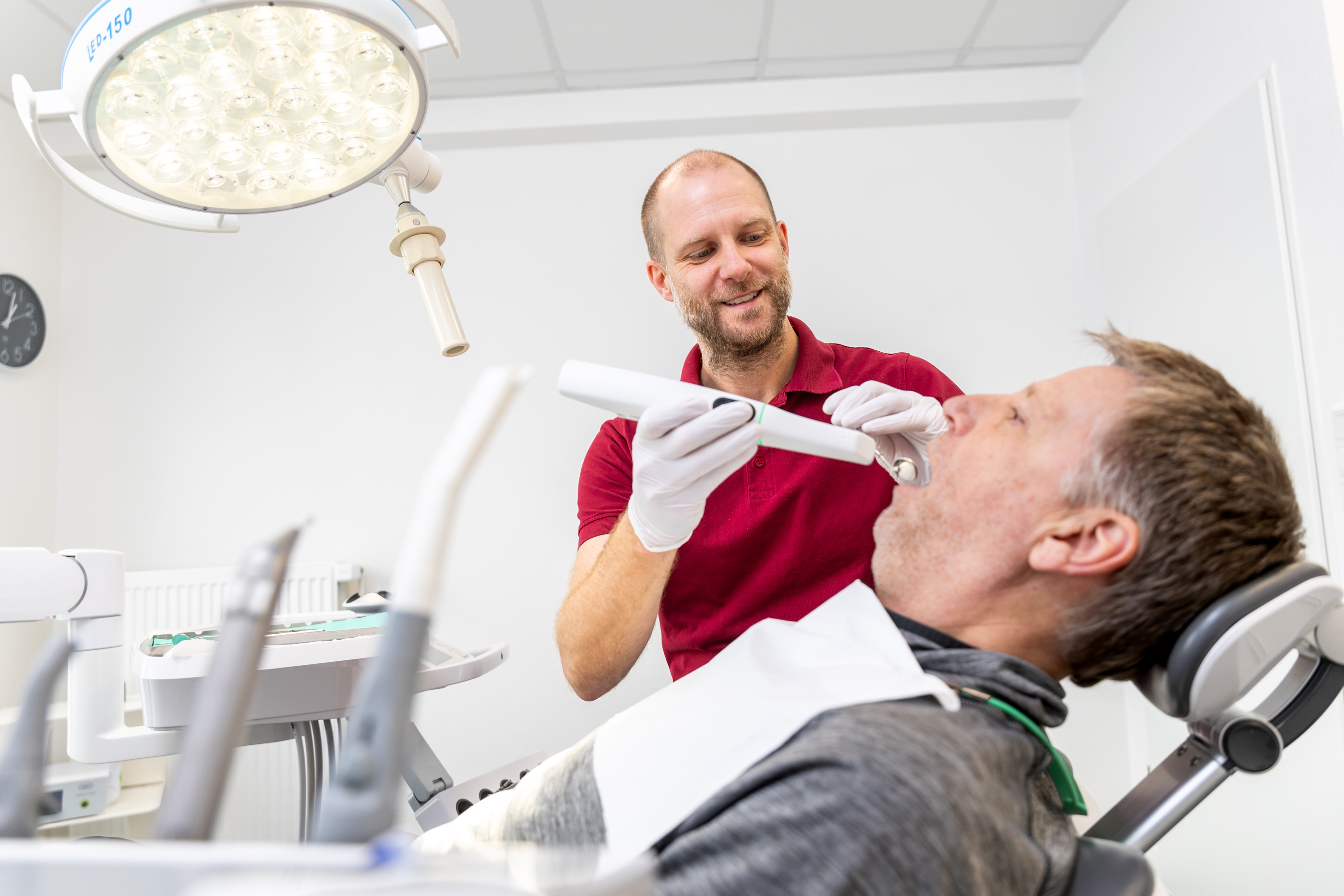 Ein Zahnarzt in einem roten Hemd untersucht mit einem Handgerät die Zähne eines männlichen Patienten auf einem Zahnarztstuhl.