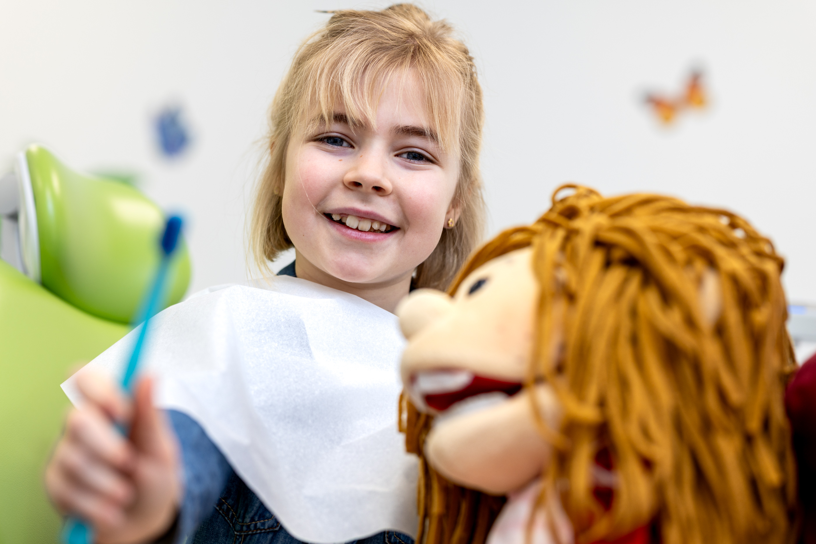 Ein lächelndes Mädchen sitzt auf einem Zahnarztstuhl und hält eine Zahnbürste und eine Puppe in der Hand.