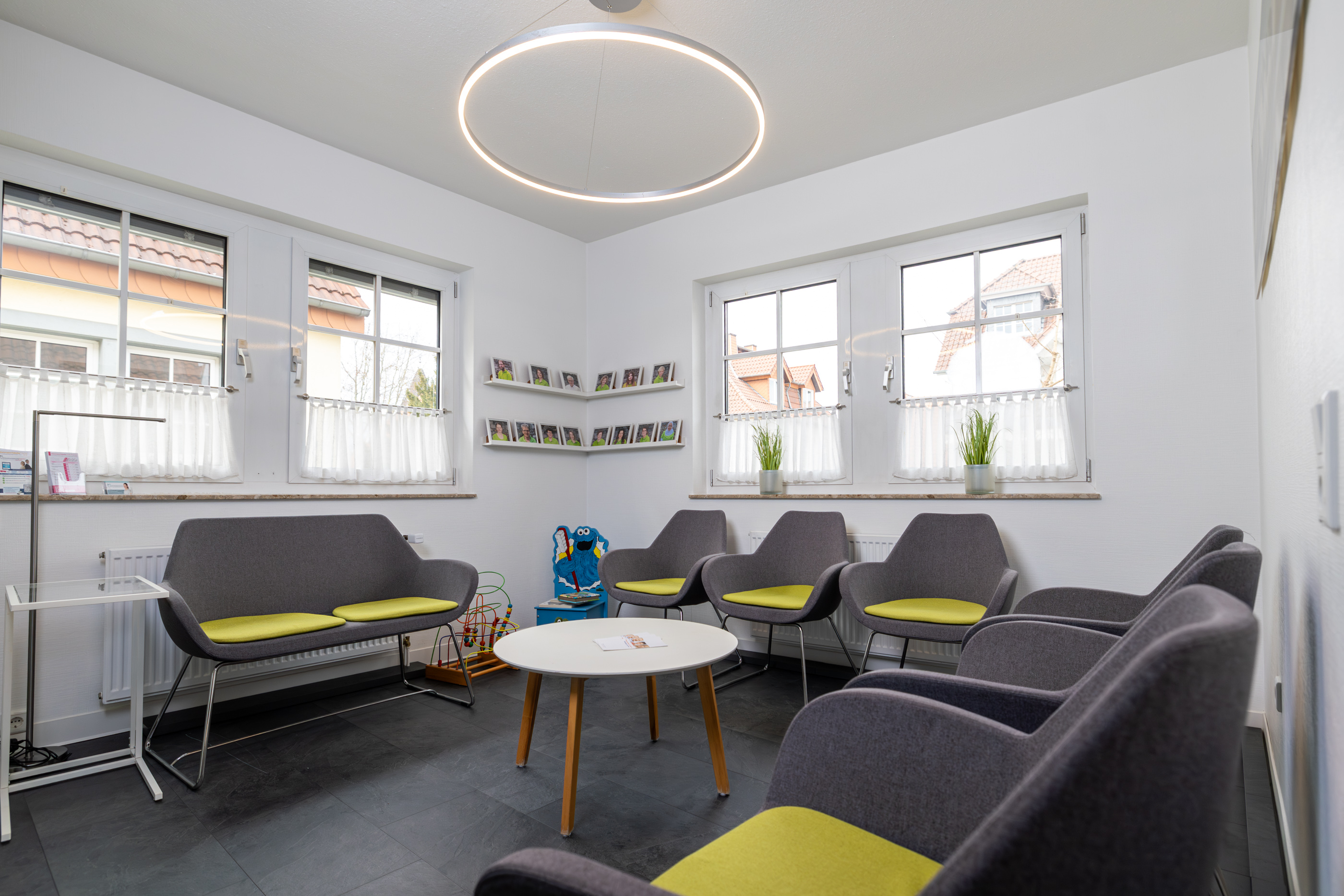 Modernes Wartezimmer mit grauen Stühlen, grünen Kissen, einem weißen runden Tisch, Fotoregalen und großen Fenstern.