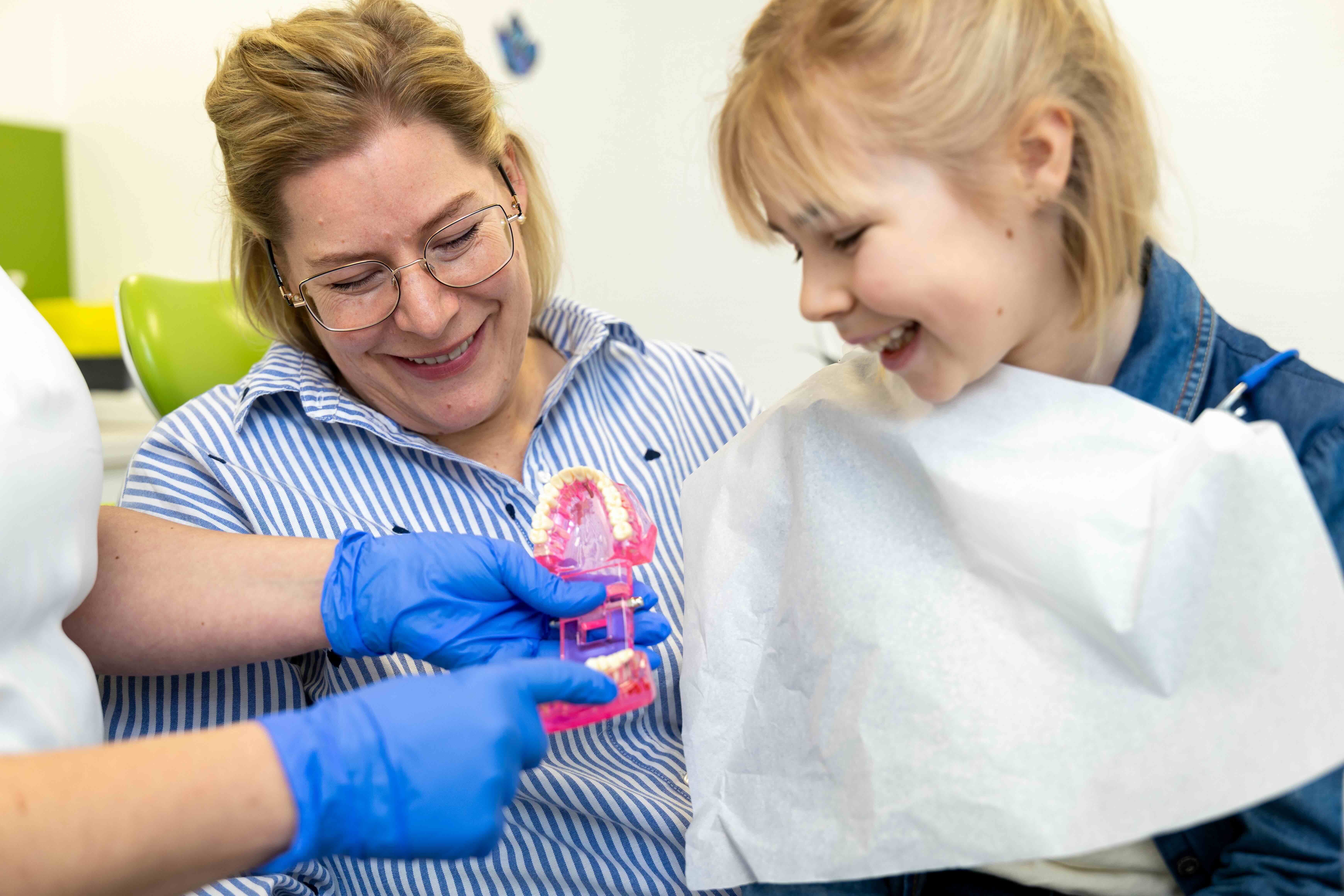 Zahnärztin mit blauen Handschuhen erklärt einer Patientin mit Zahnbehandlungsdecke an einem Zahnmodell die Mundpflege.
