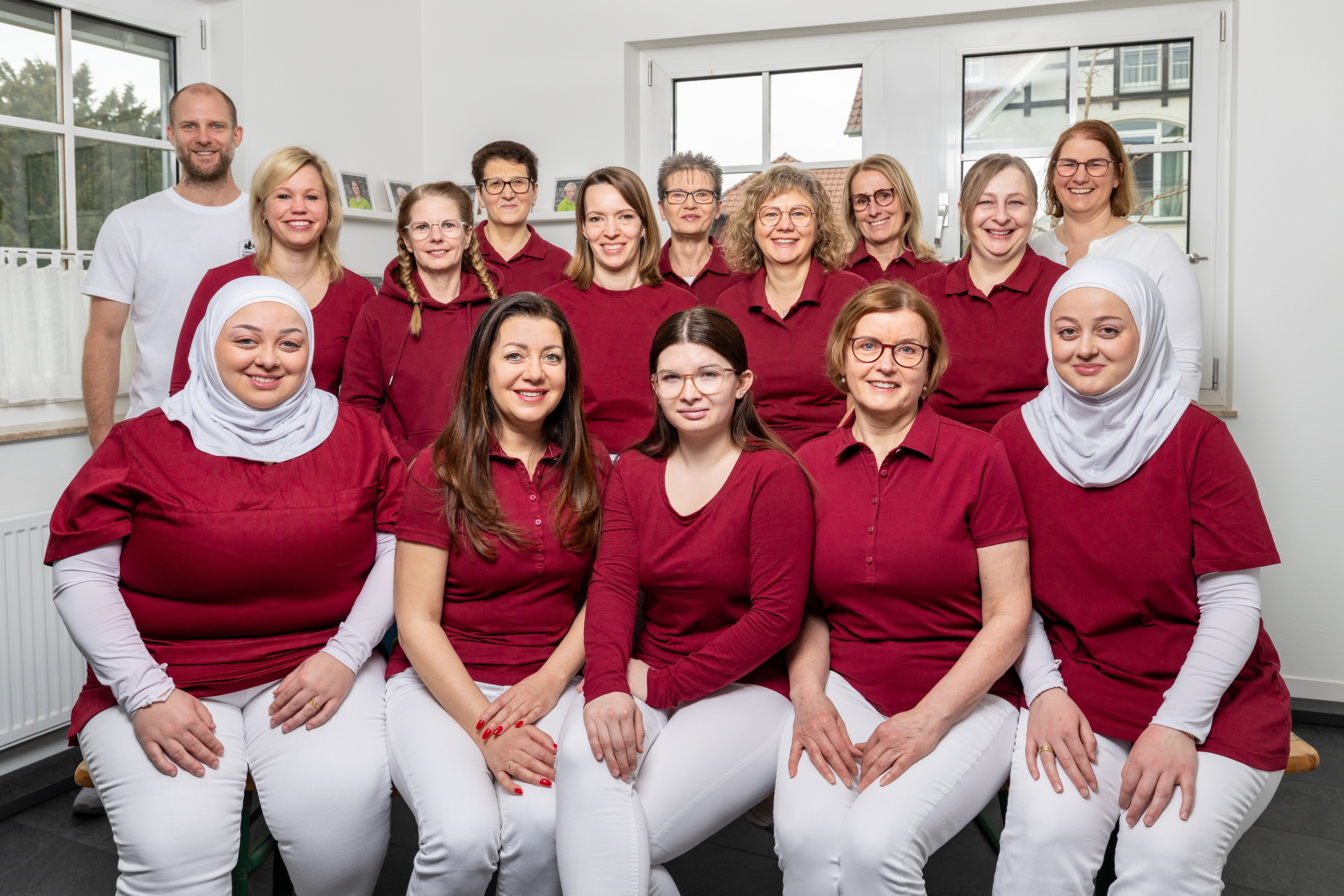 Gruppenbild eines Teams in einem hellen Raum mit Fenstern. Das Team besteht aus 16 Personen, 14 Frauen und 2 Männern. Die meisten Frauen tragen rote oder bordeauxfarbene Oberteile und weiße Hosen, zwei der Frauen tragen weiße Kopftücher. Die beiden Männer tragen weiße T-Shirts. Die Personen sitzen und stehen in drei Reihen und lächeln in die Kamera.