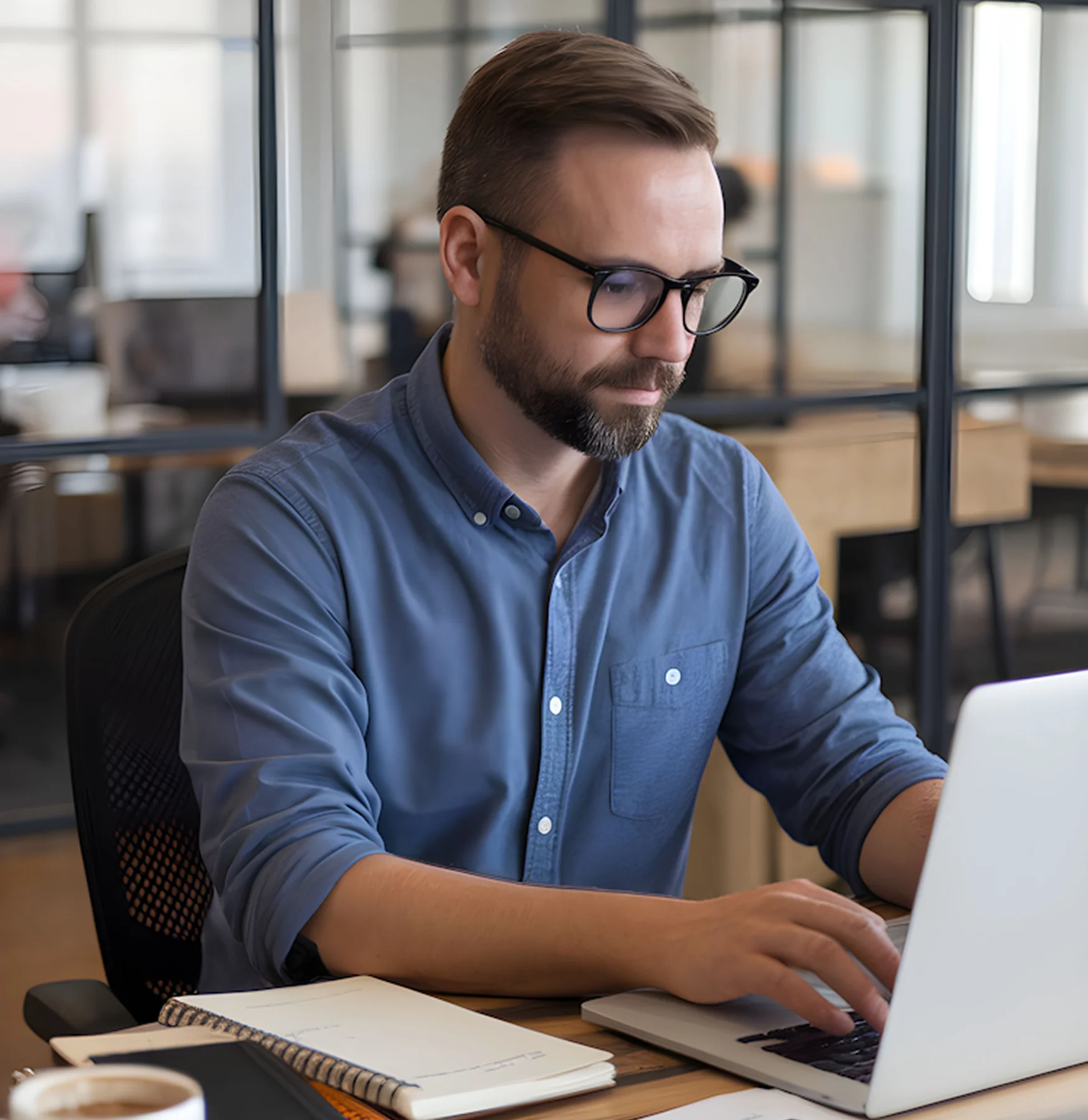 Man with glasses and beard working on a laptop in a modern office setting.