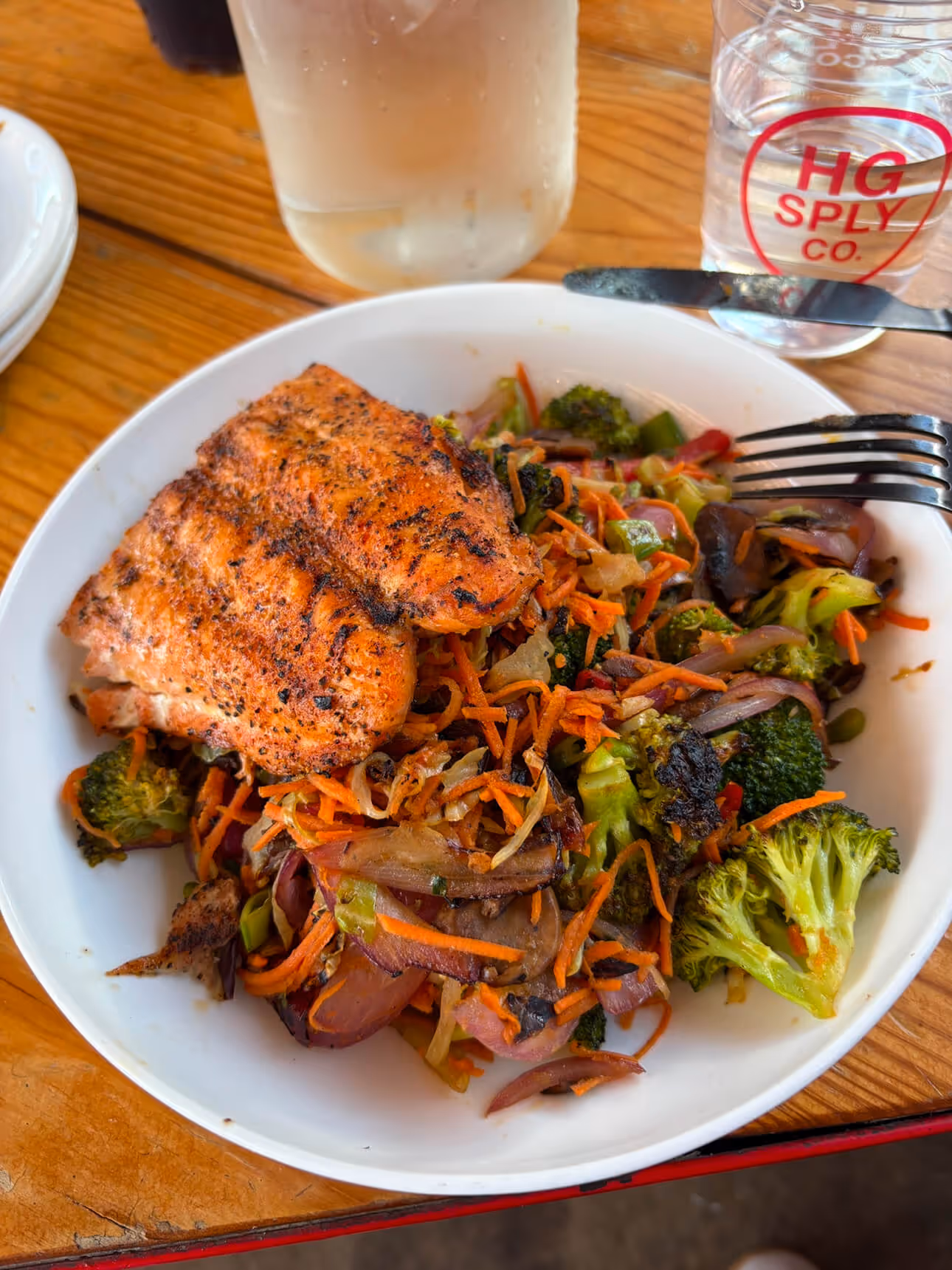 Grilled salmon served with sautéed broccoli, carrots, onions, and peppers in a white bowl on a wood table.