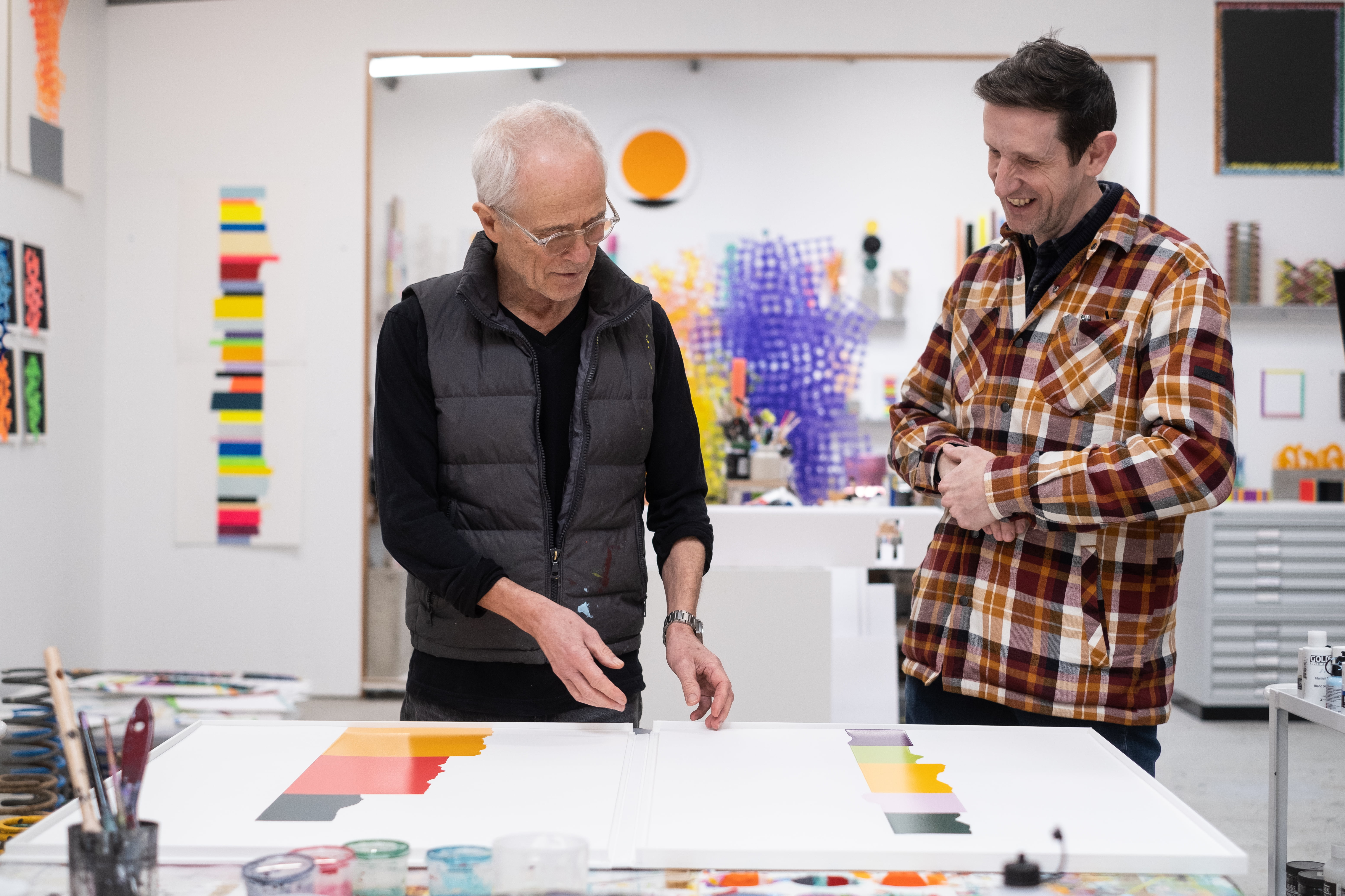 David Batchelor and master printer Peter Bennett studying two Chromophilia prints on a studio table, colourful artworks and materials filling the room behind them.