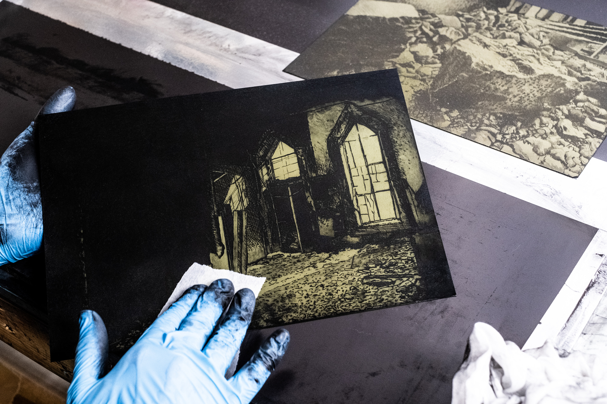 Wiping ink from the Mosul Museum III etching plate, gloved hand and rag, further inked plates on the bench.