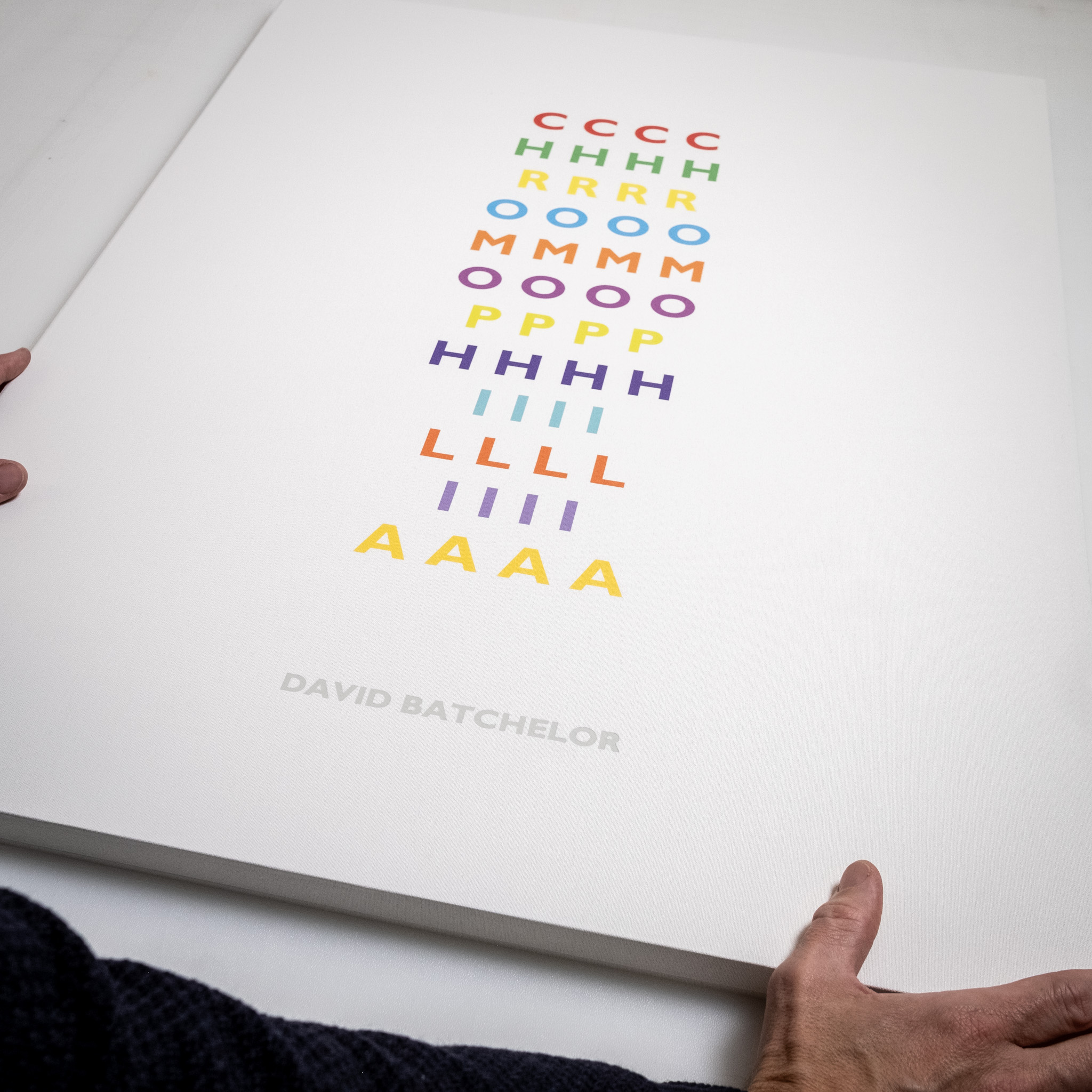 Hands holding a the Chromophilia box set flat, showing the stacked coloured lettering and its scale against a white surface.