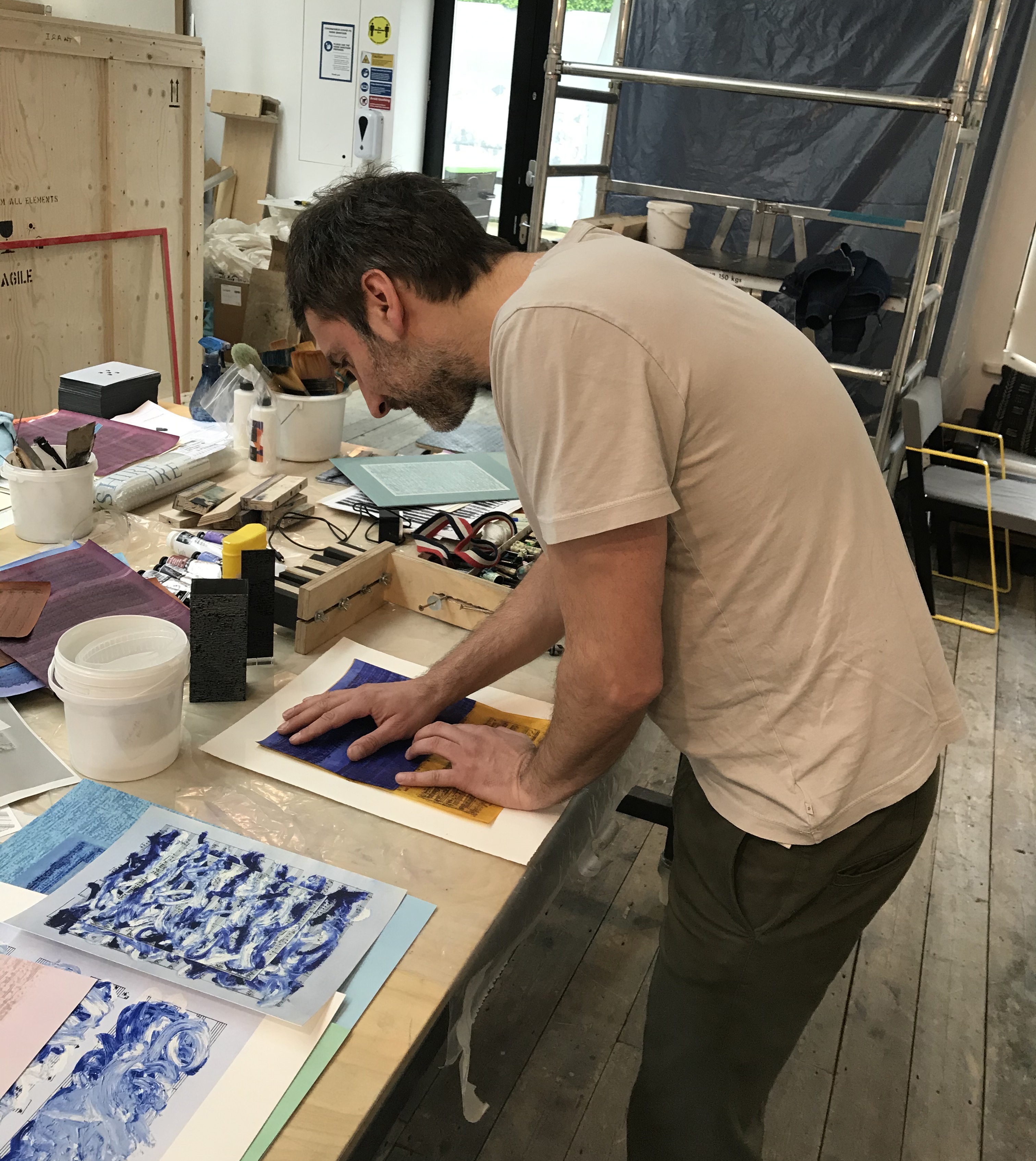 Idris Khan in the studio at Atelier JI, pressing a blue and yellow score-layered sheet onto paper, proofs spread on the table.