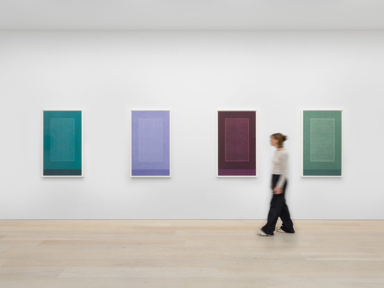 Four large framed prints in teal, periwinkle, wine-red and sage green hang evenly on a white gallery wall, each with a rectangular text field; a visitor walks past.