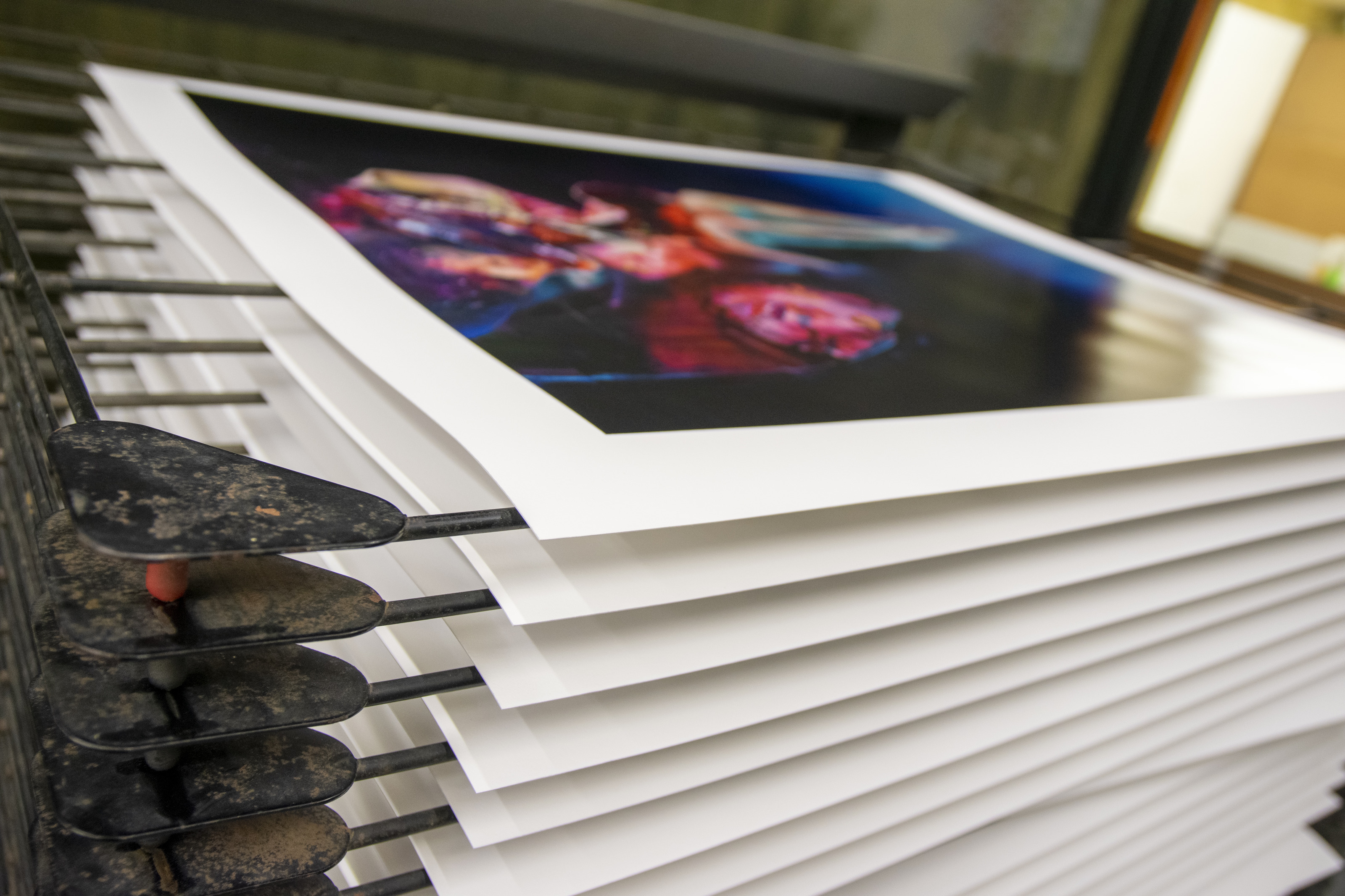 Freshly printed Chloe Early editions stacked on a studio drying rack at Atelier JI, the top sheet showing a jewel-toned figurative image.