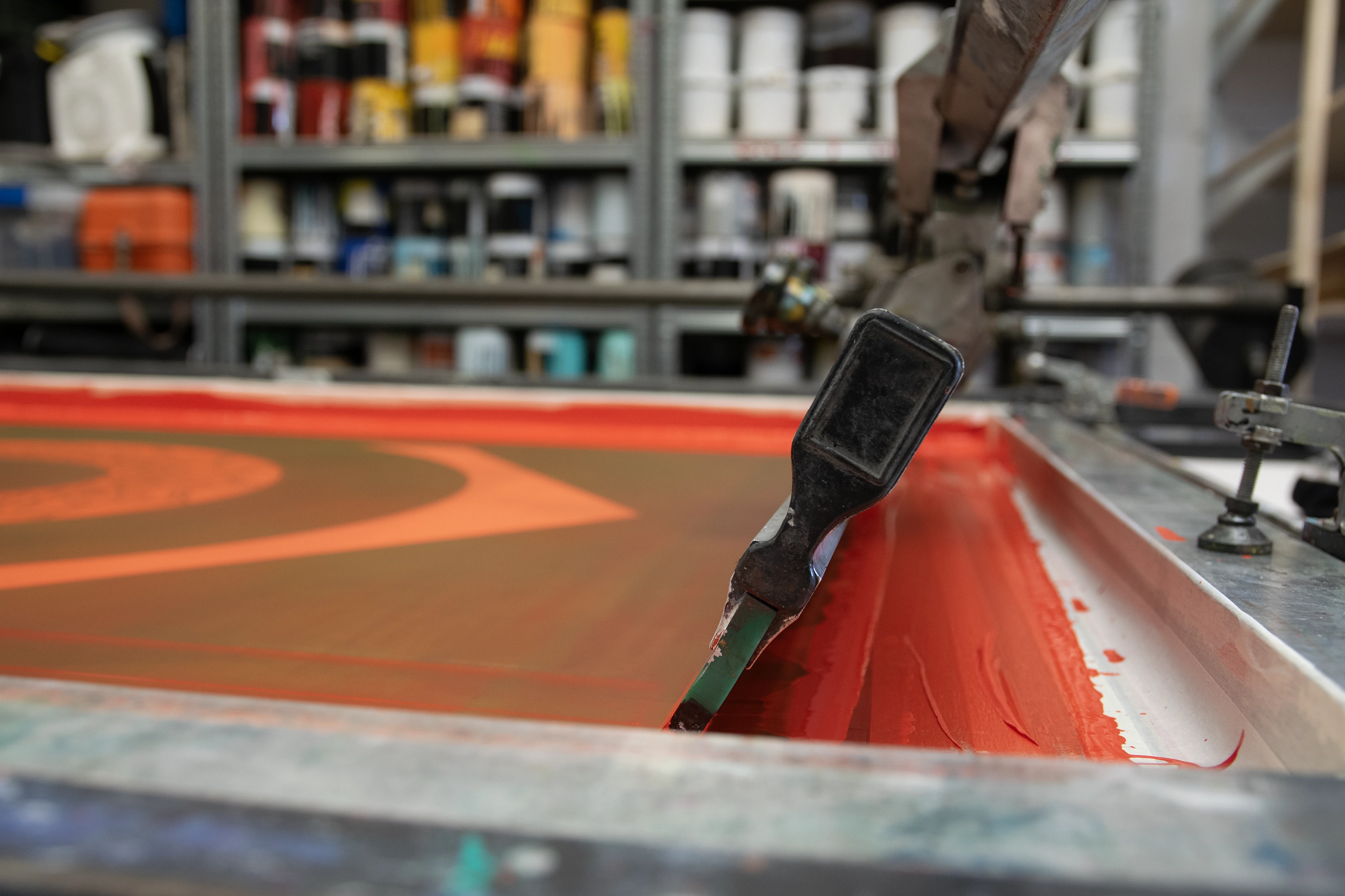 A squeegee resting on a screen bed at Atelier JI, a bead of red-orange ink along its edge above the Fairey dove print.