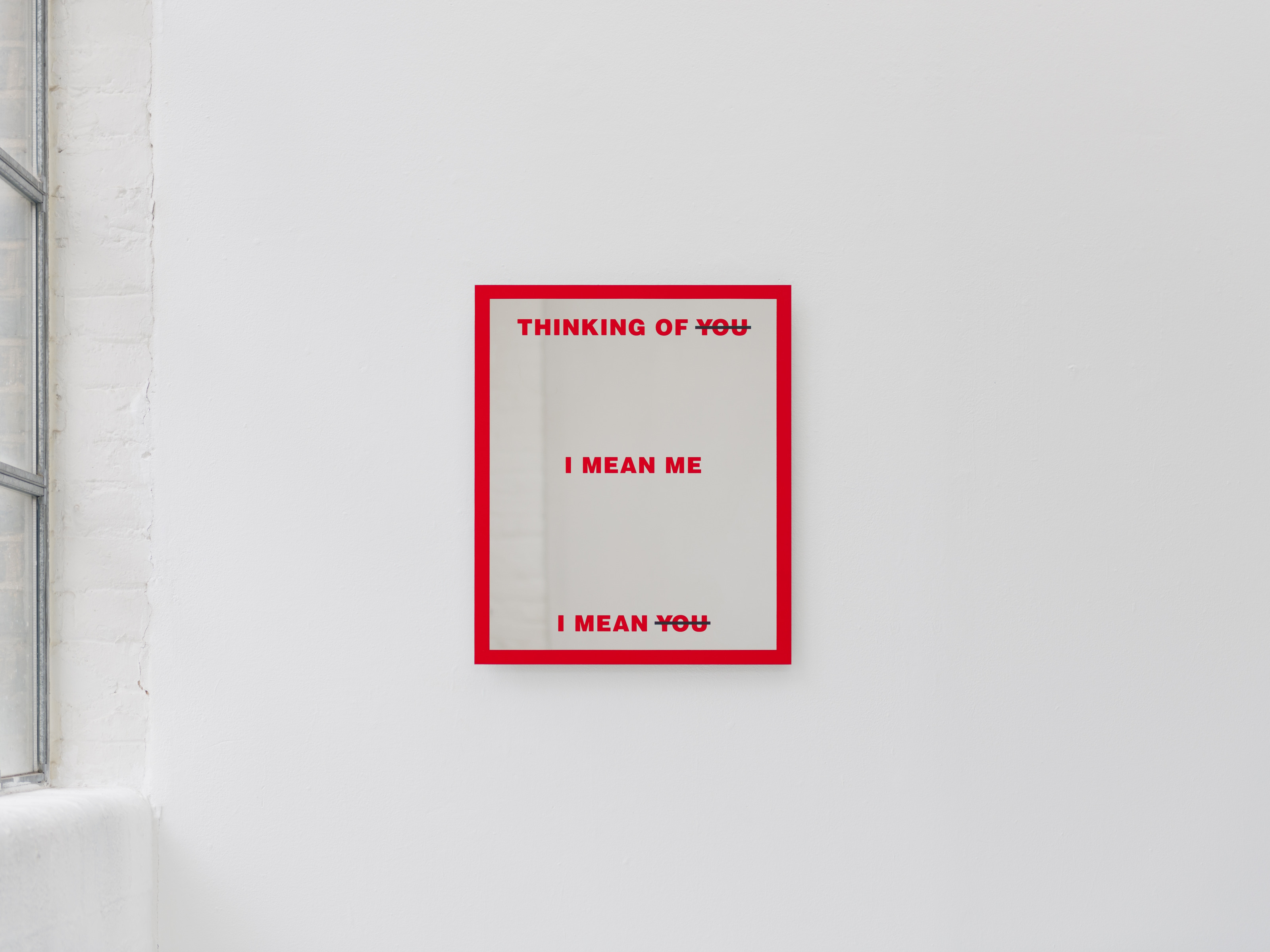 Barbara Kruger's Untitled (Mirror) hangs on a white gallery wall beside a window, the red-bordered mirror reflecting the room.