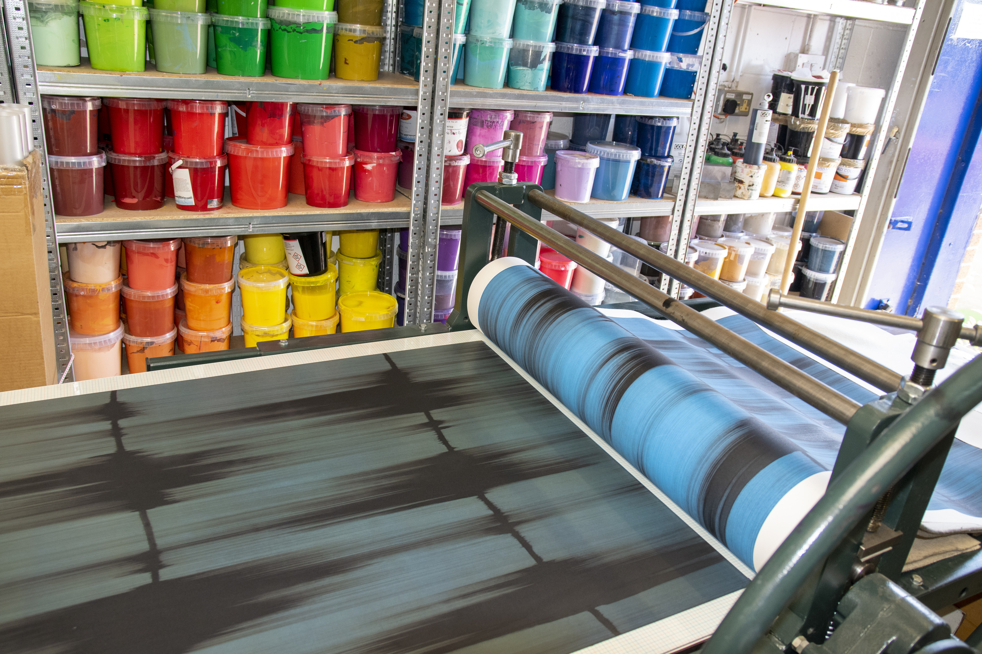 Mark Francis's blue 'Echo 2' being printed on the roller press at the Atelier JI studio, tubs of coloured ink shelved behind.