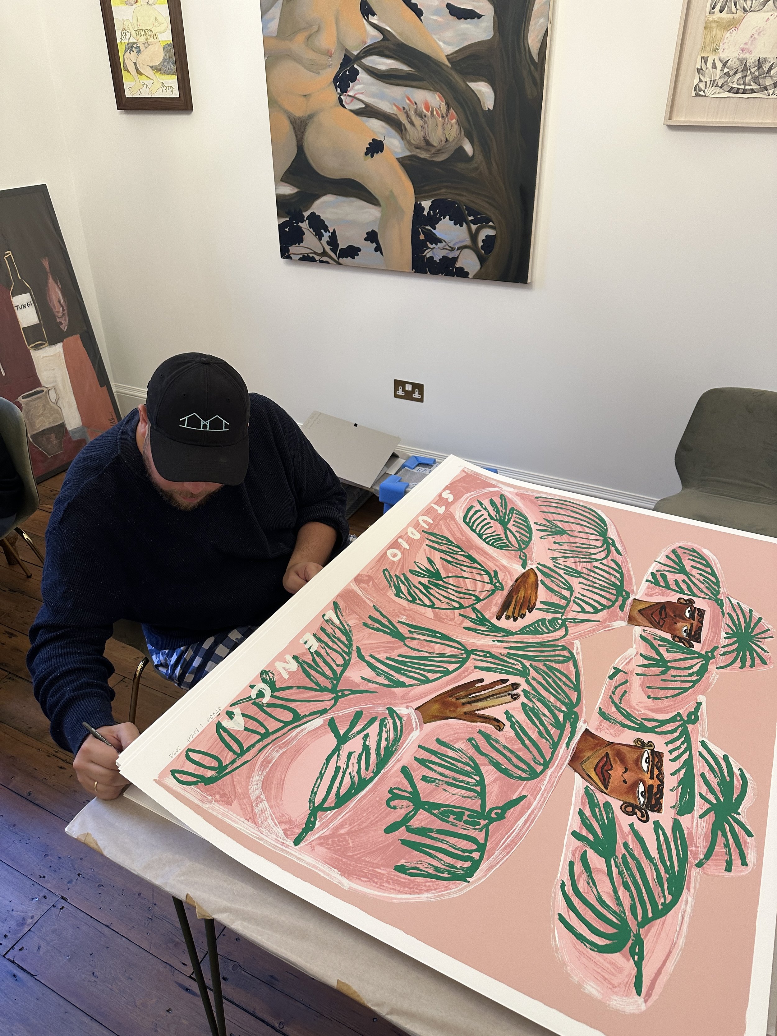 Studio Lenca (José Campos) signing a Lora print at a table, his own paintings hung on the wall behind him.
