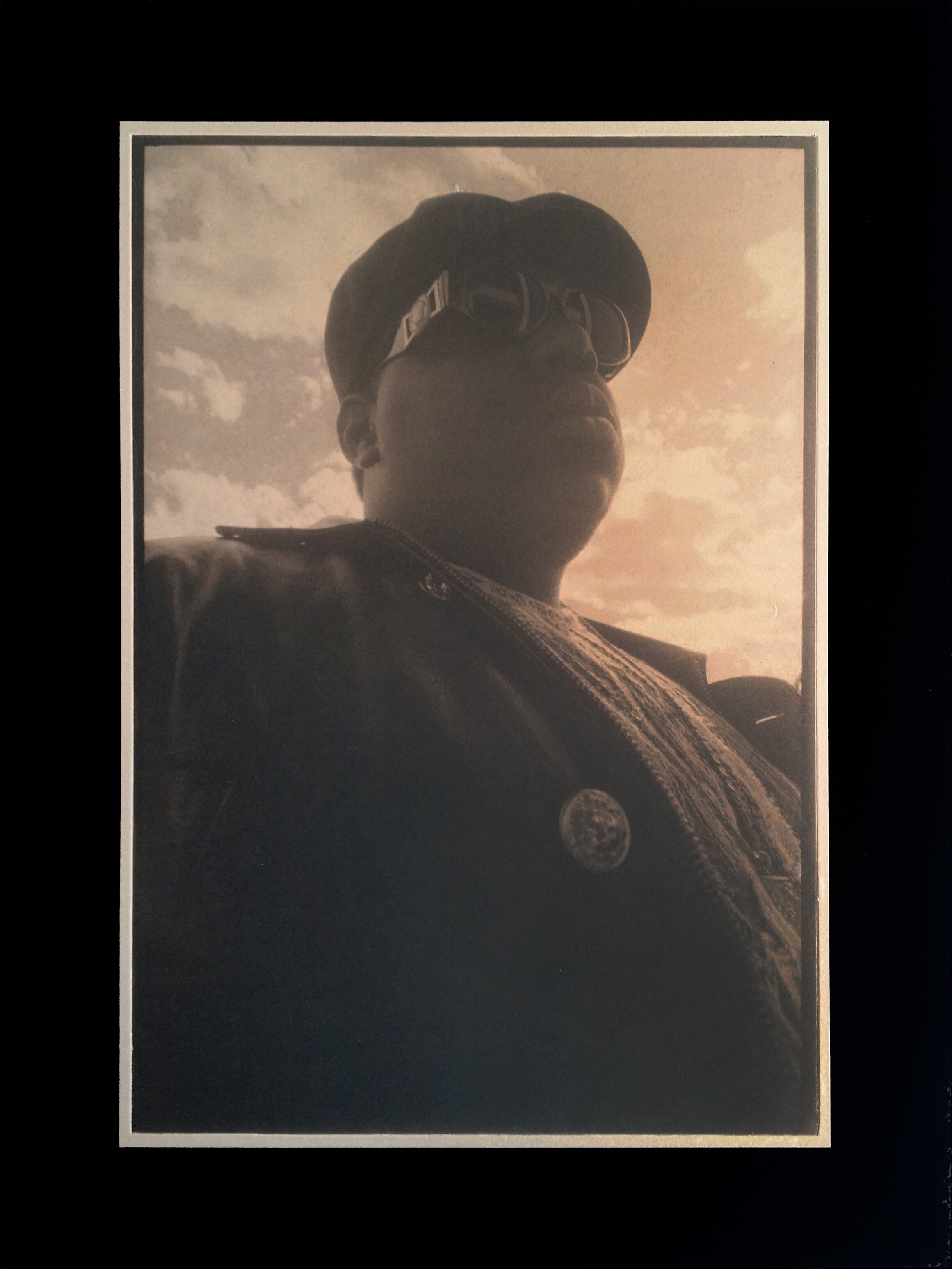 Eddie Otchere's 'Biggie Smalls': a low-angle sepia-toned portrait of the rapper in a flat cap and shades against a cloudy sky.