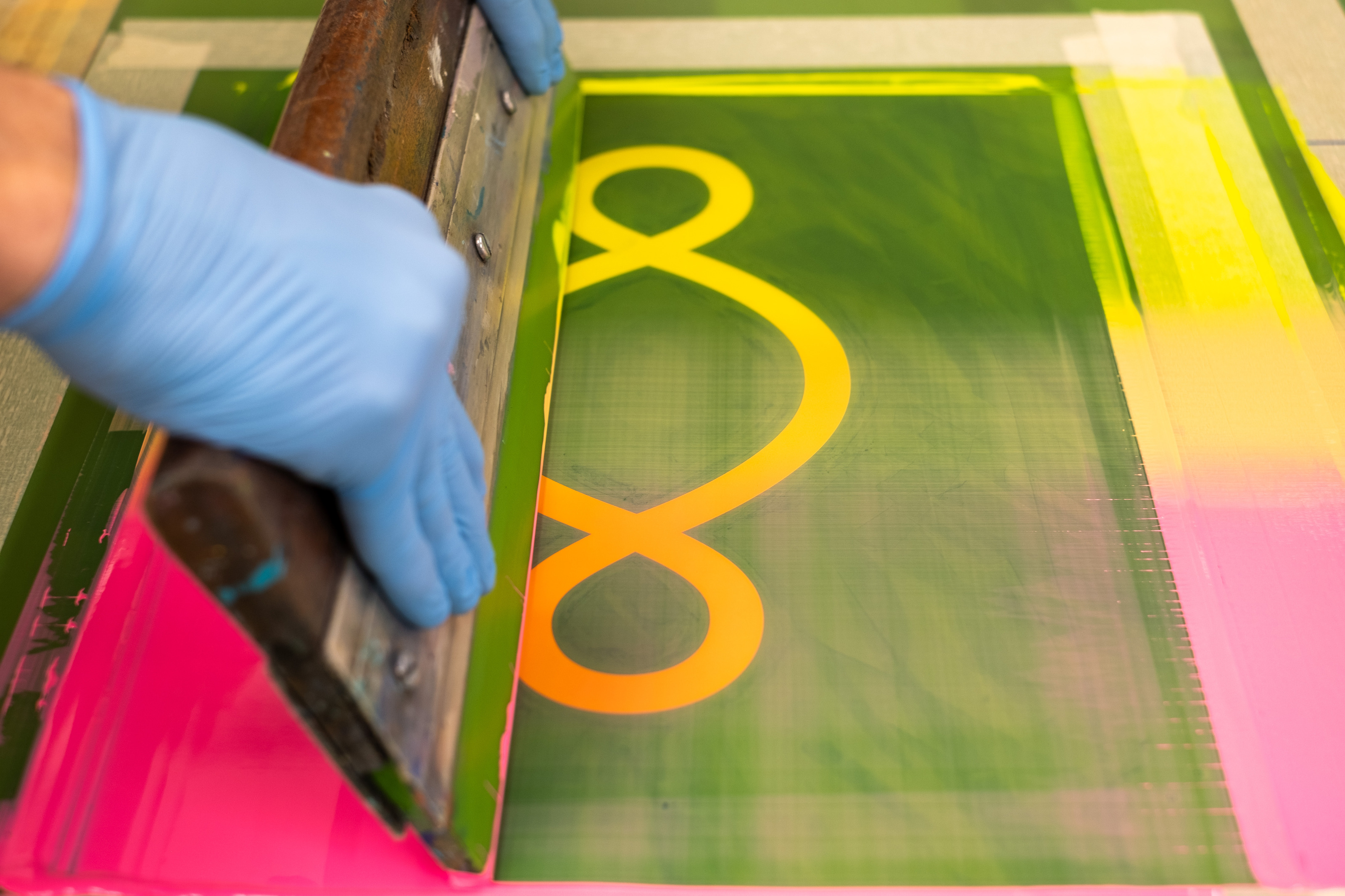 Printing Pistoletto’s Preventive Peace Flags at Atelier JI: a gloved hand pulls a squeegee of blended pink and yellow ink across the screen