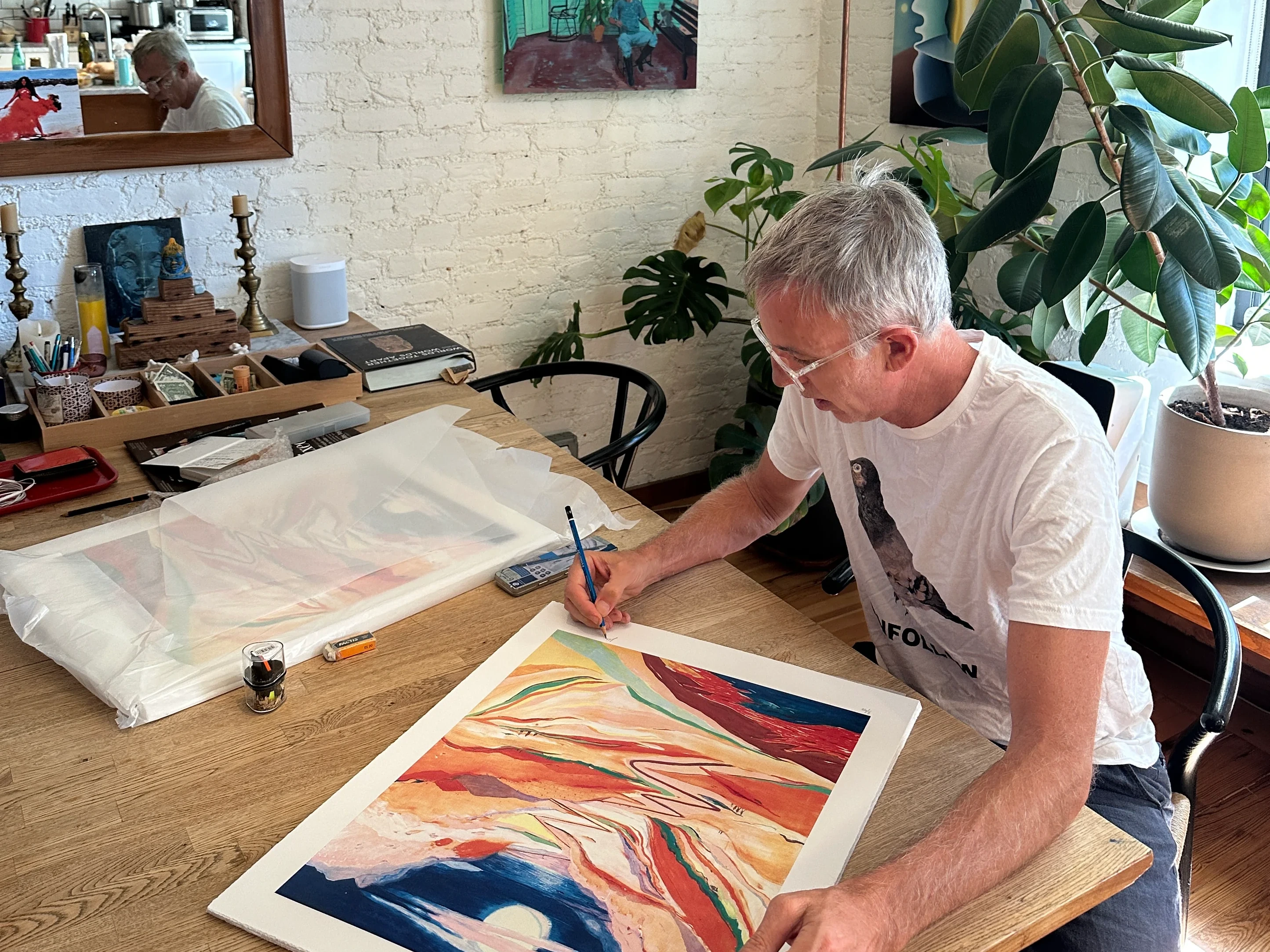 Jules de Balincourt signs an impression of They Come to Get Lost in pencil at a wooden table in his plant-filled studio